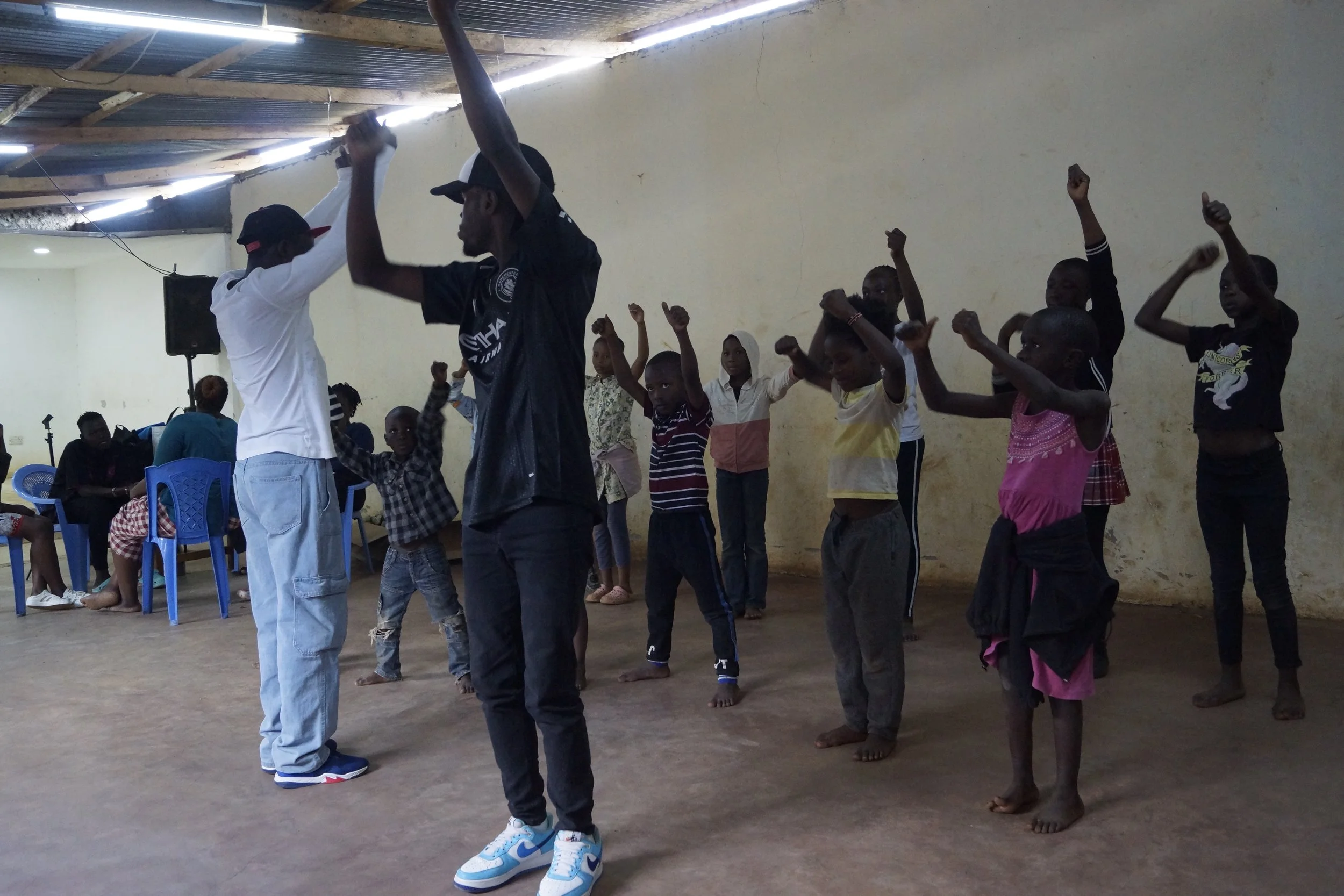 Children participating in an indoor activity with two instructors, raising their arms, some barefoot, in a spacious room with a high ceiling and a few chairs in the background.