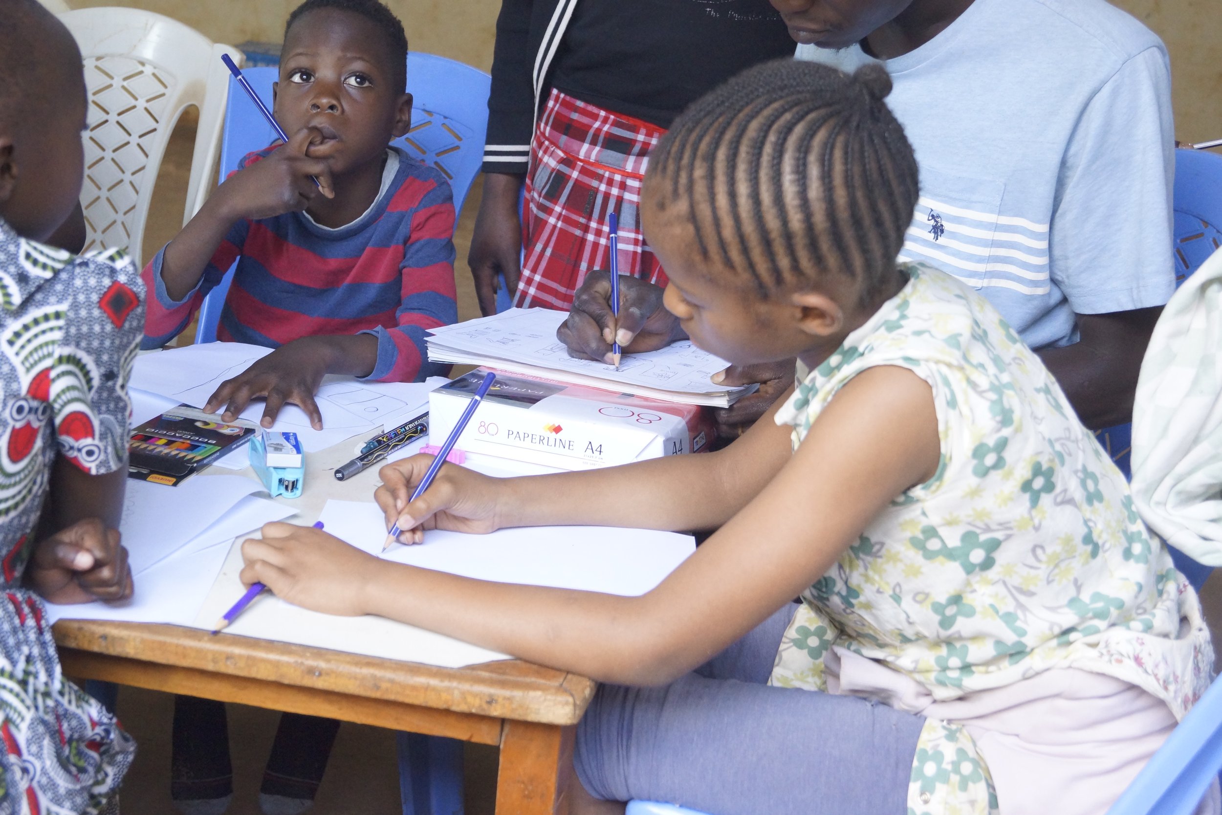 Children and a young woman engaged in drawing or writing activity at a wooden table in a classroom setting, with supplies like paper, pens, and markers.