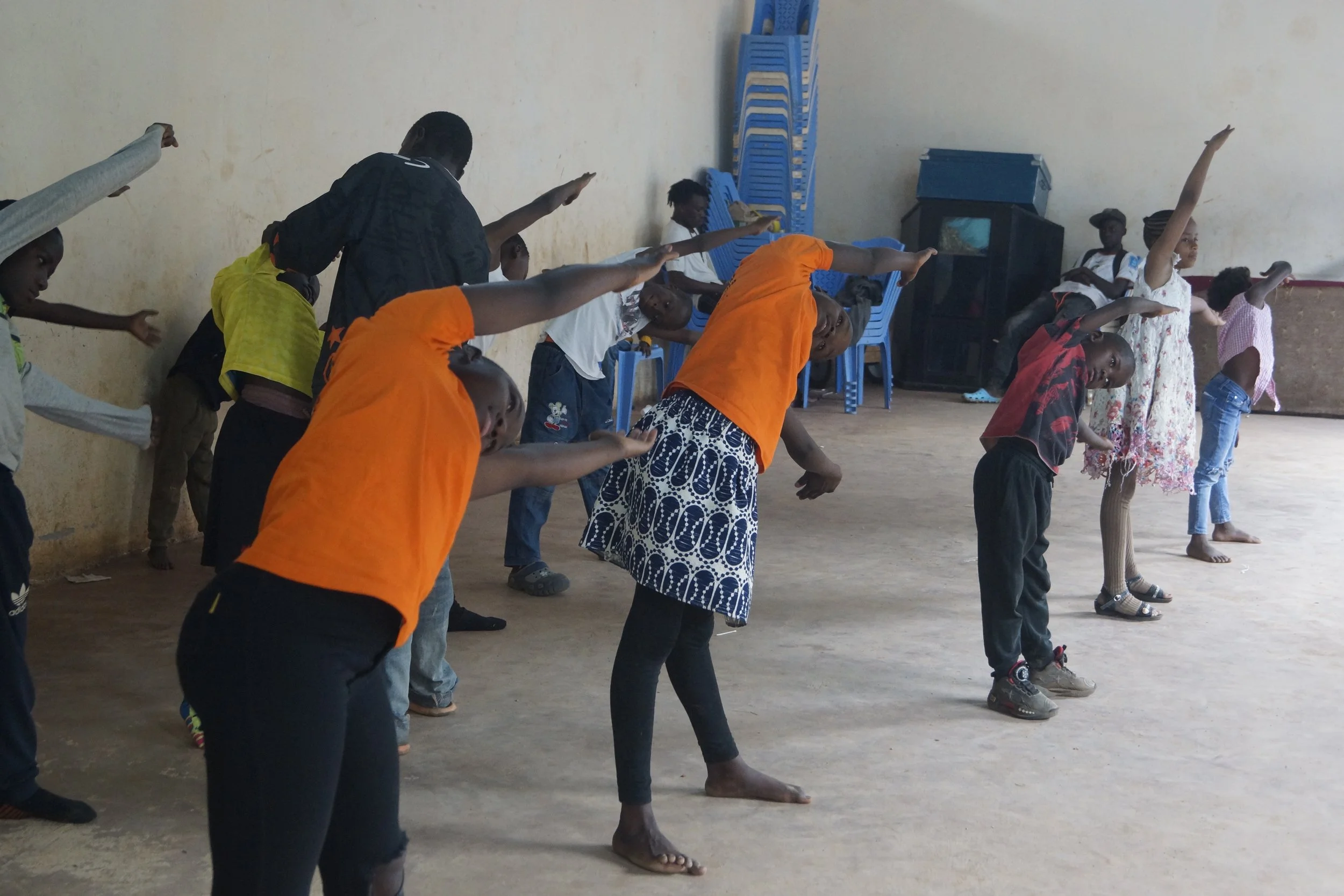 Children participating in a group activity or exercise in an indoor space, some are stretching or bending to the side, with chairs and storage cabinets in the background.