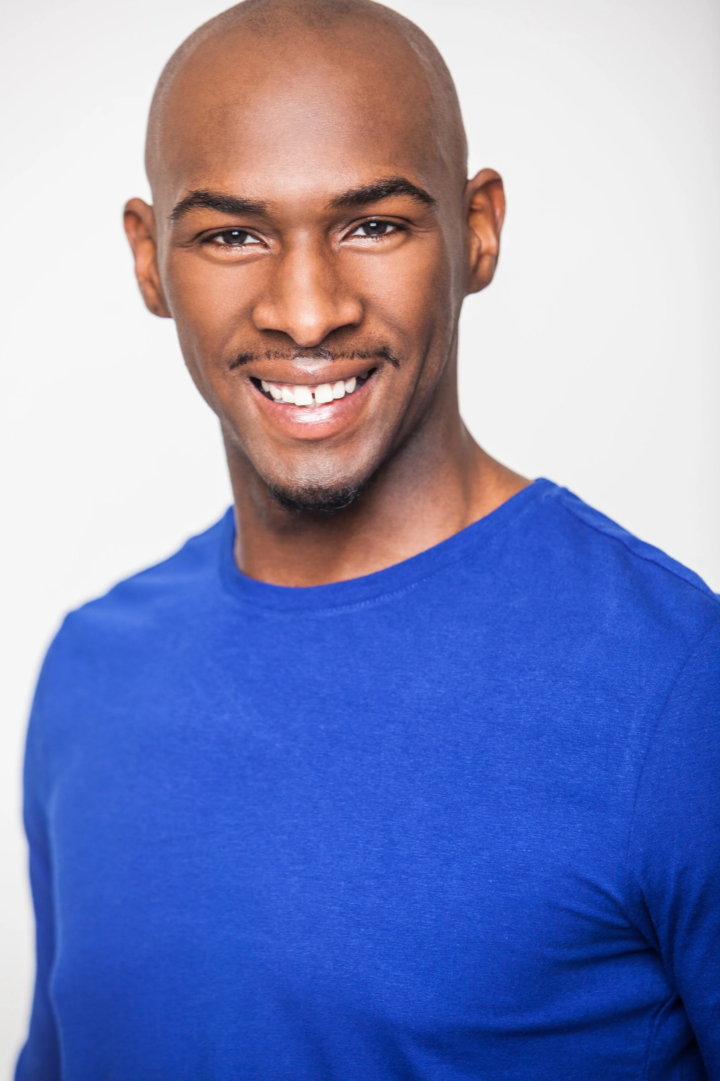 Smiling young man with a shaved head wearing a blue t-shirt against a white background.
