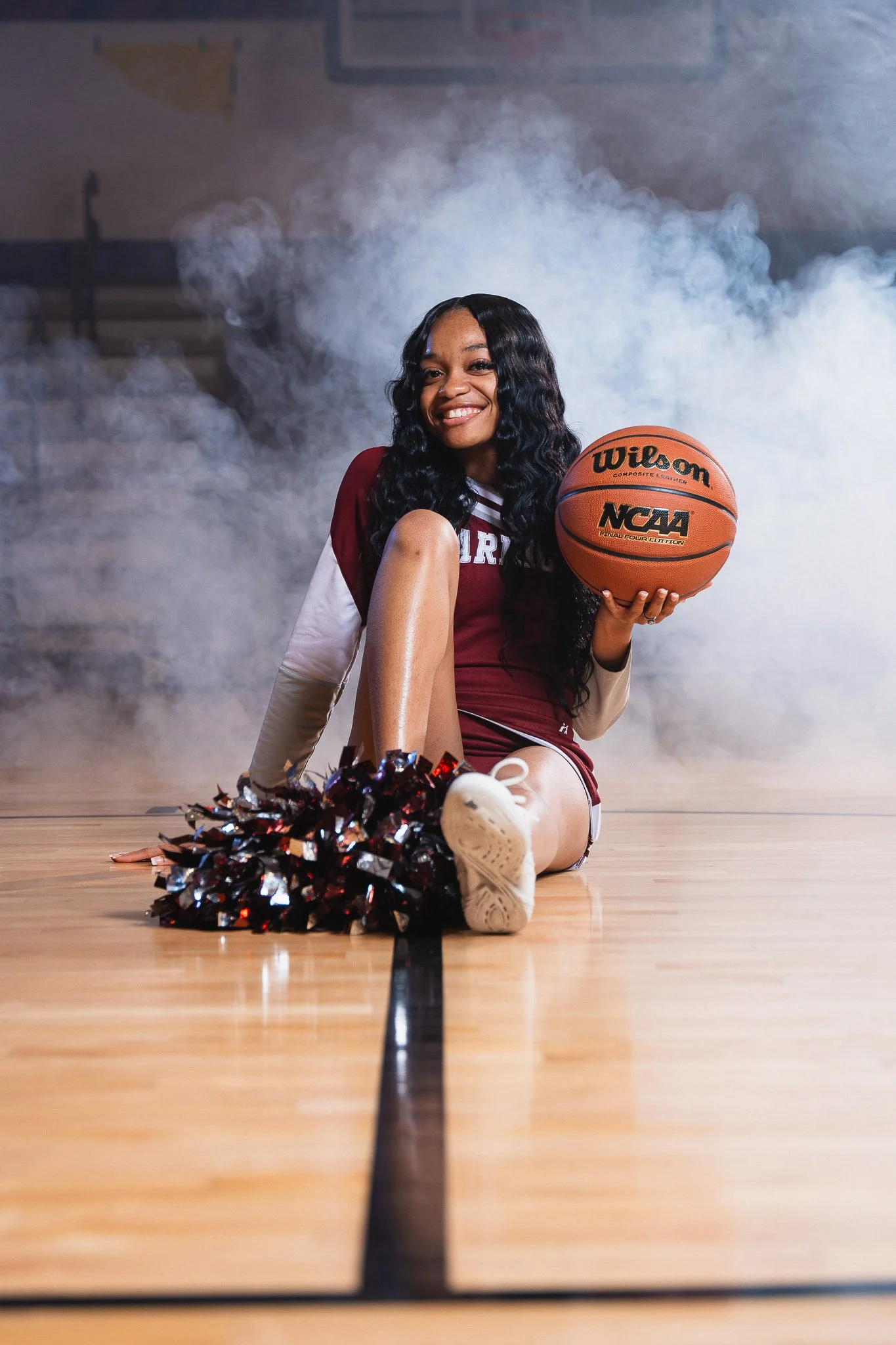 A female basketball player sitting on the court with a basketball in her hand, smiling, with a cheerleading pom-pom in front of her, wearing a maroon and white uniform, and surrounded by smoke.