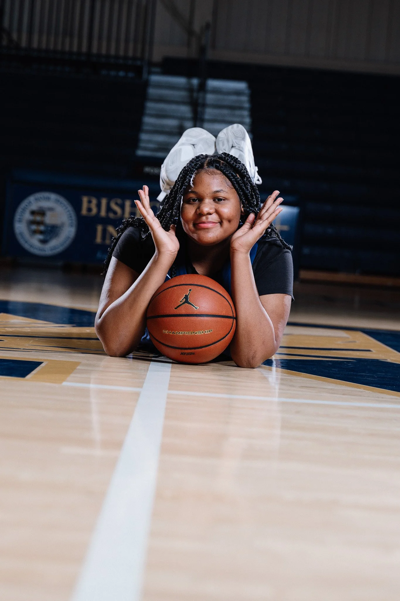 A young girl lying on a basketball court with her head resting on a basketball, smiling at the camera. She is wearing black clothing and has braided hair. The court has blue and yellow markings, and there is a basketball hoop visible in the backgroun