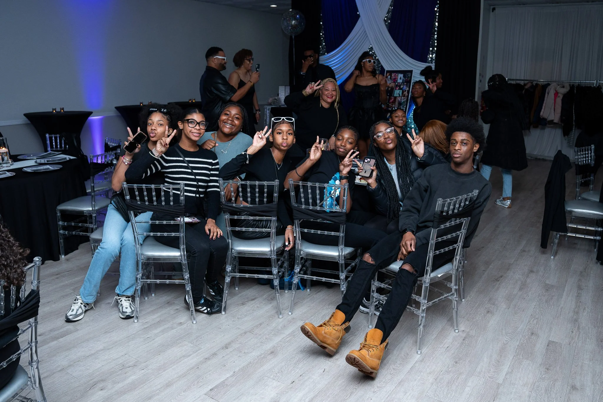 Group of young people sitting and standing together at a party, many making peace signs and smiling. The setting has tables, chairs, and decorative curtains with blue and white draping.