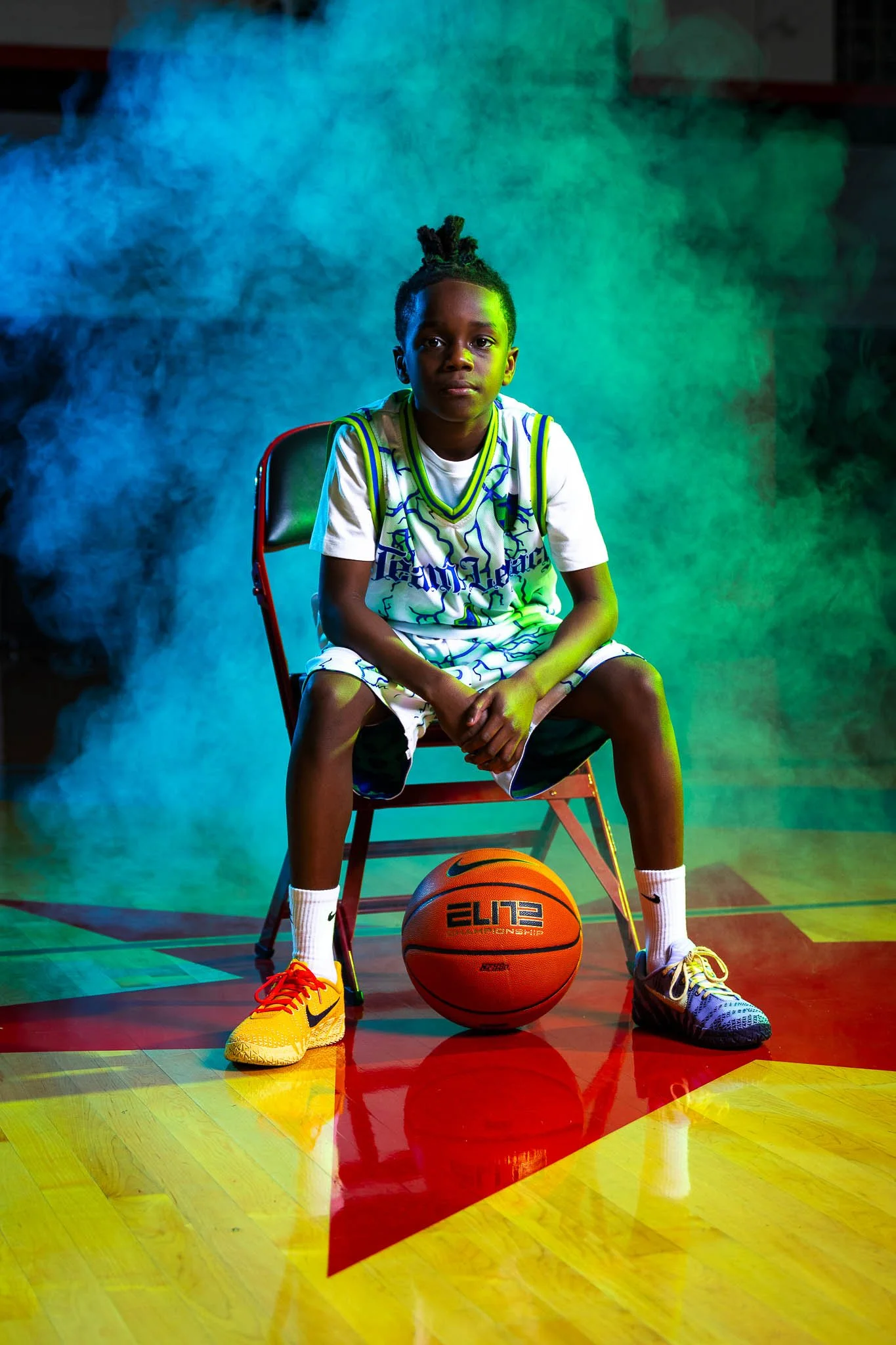 Young boy sitting on a basketball court with a basketball in front of him, wearing sports shorts, a basketball jersey, sneakers, and white socks, with colorful lighting and smoke effects in the background.