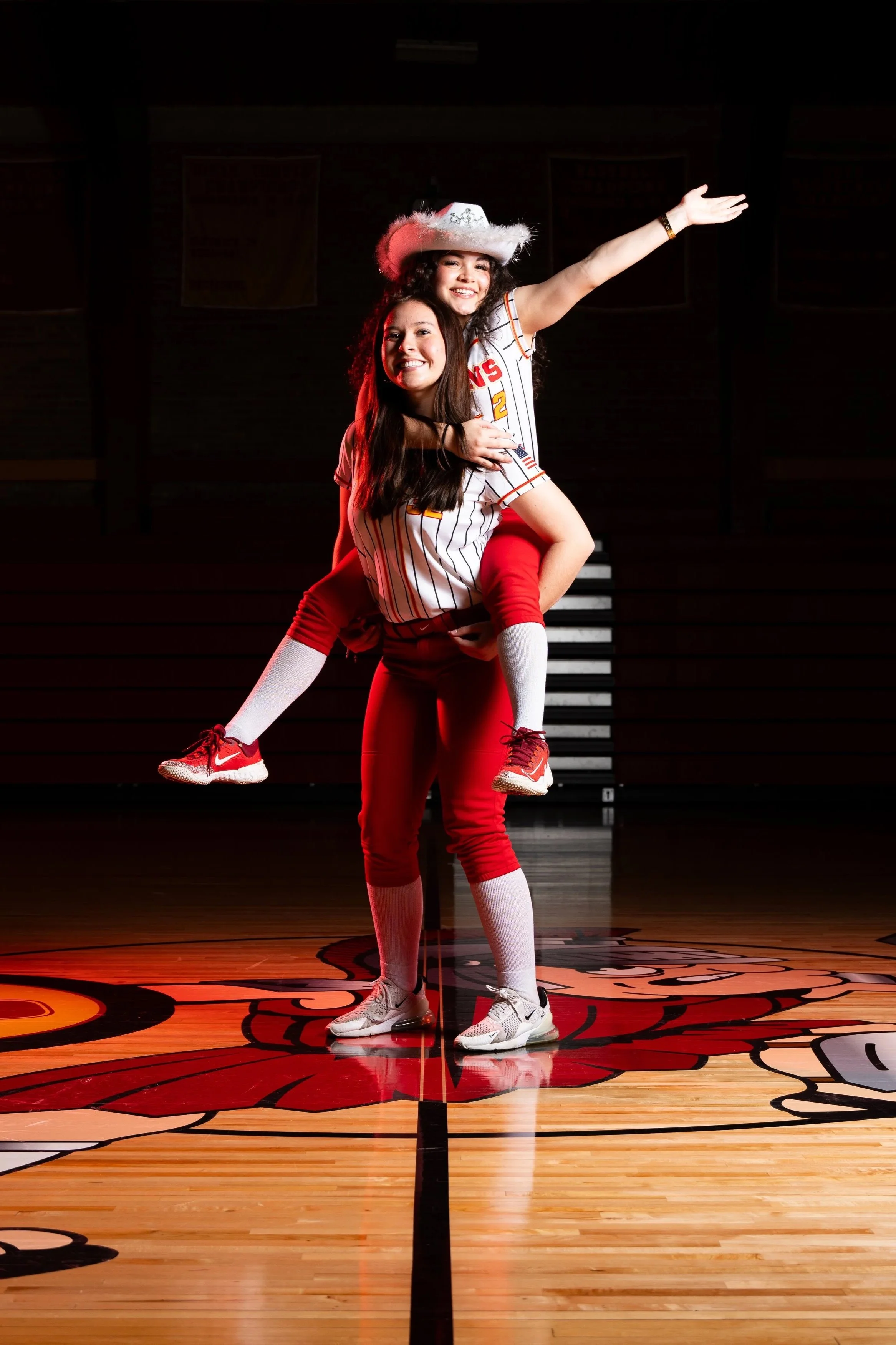 Two young women in basketball uniforms, one sitting on the other's shoulders, posing cheerfully on a basketball court with a team logo painted on the floor.