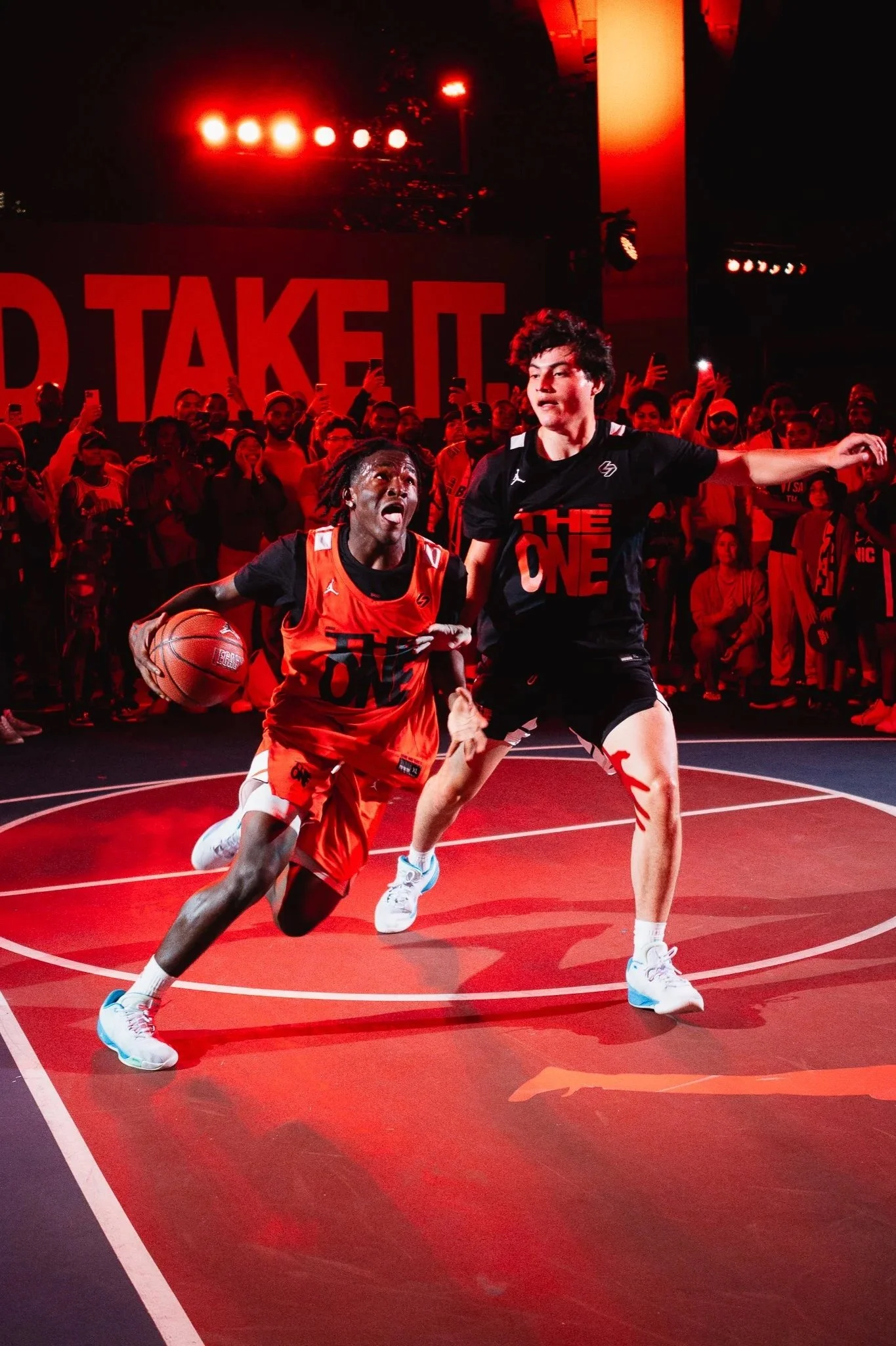 Two young male basketball players compete on an indoor court during a game or event, with an audience watching in the background. Red lighting and large signage reading 'THE ONE' are visible.
