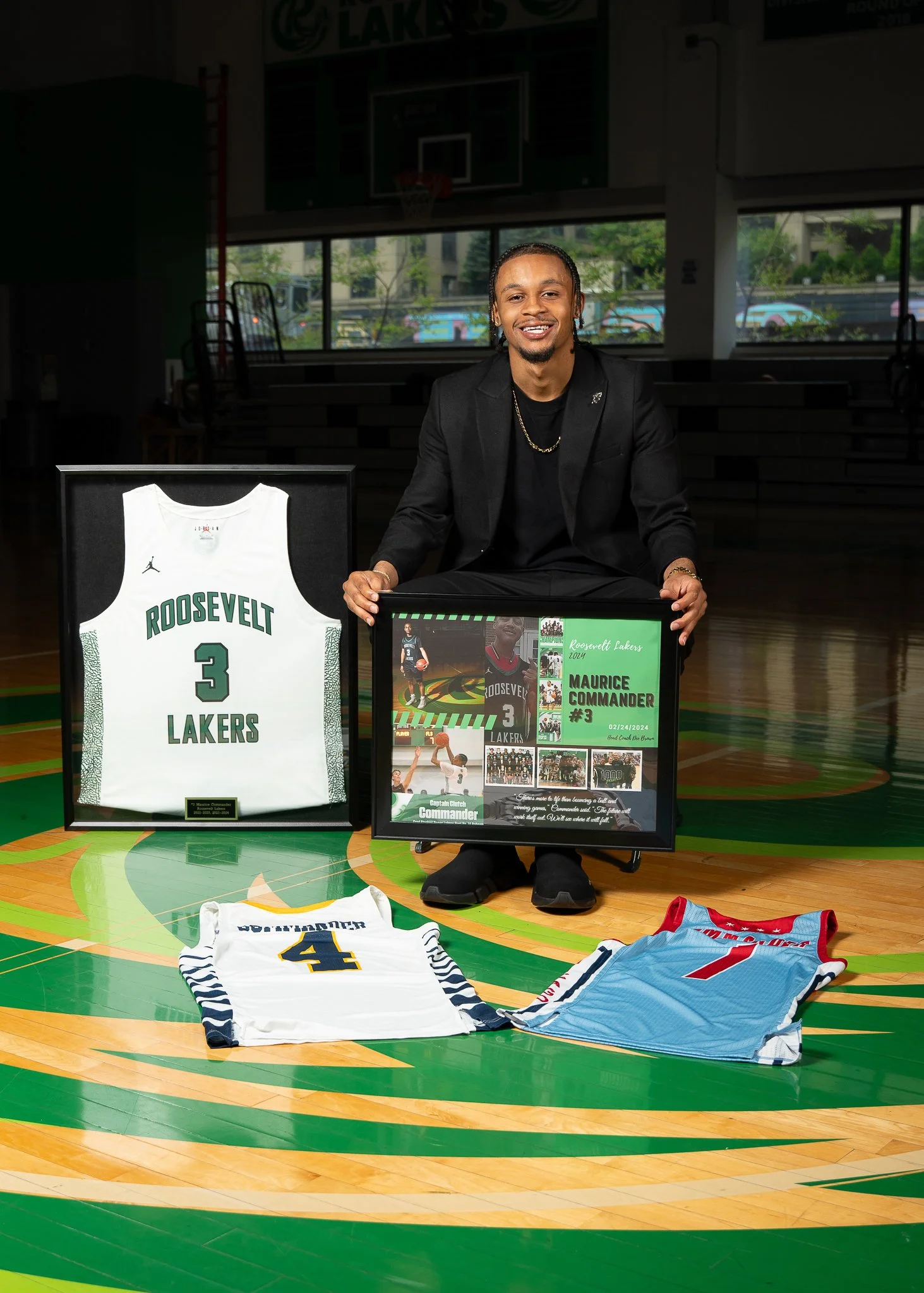 A man in a black suit kneeling on a basketball court, holding a framed collage of a basketball player, with a framed Lakers jersey and three other jerseys laid on the court in front of him. There is a large Lakers jersey displayed to his left.