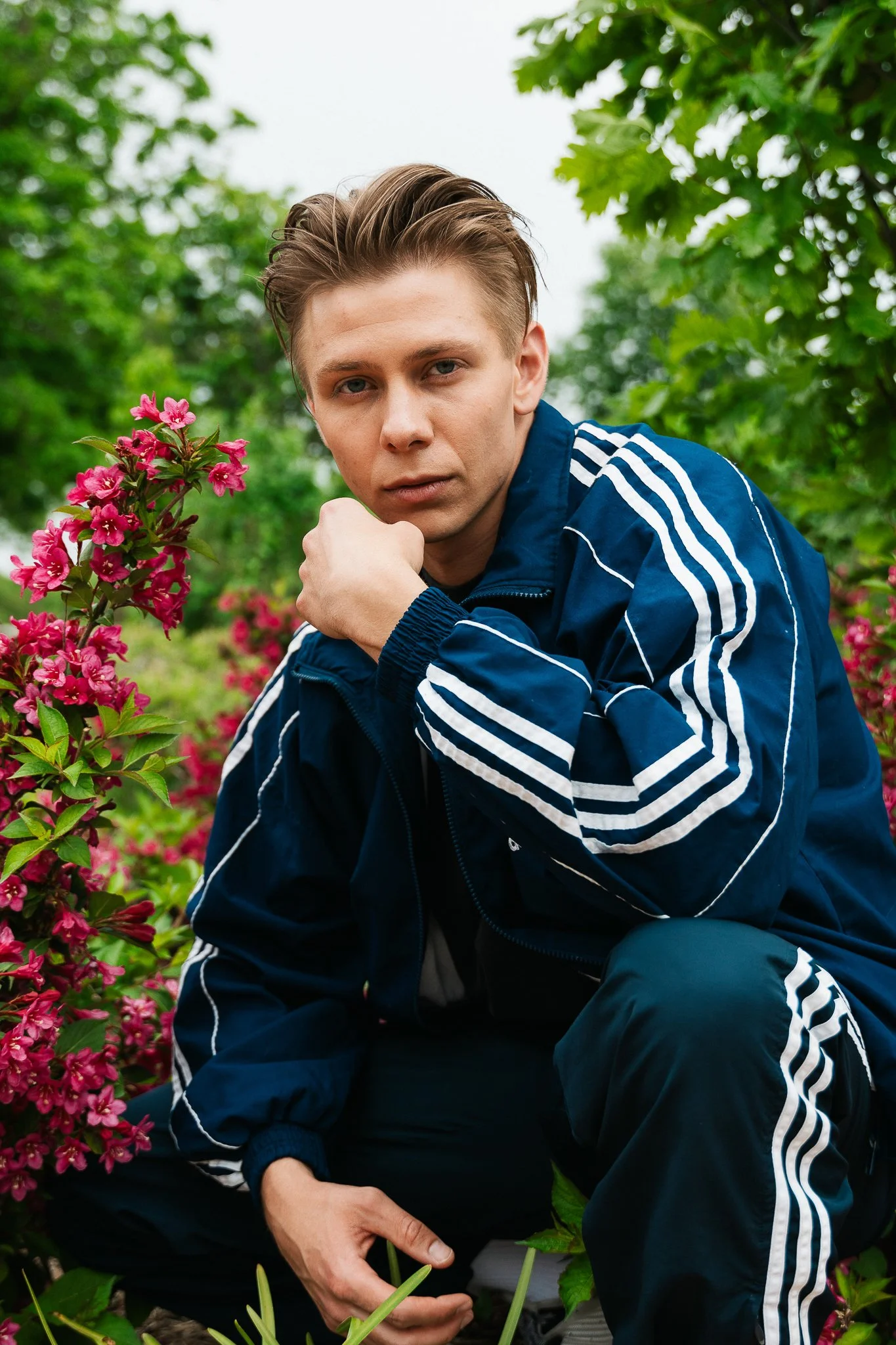 A young man in a blue athletic tracksuit crouches in a garden with pink flowers and green foliage, looking at the camera.