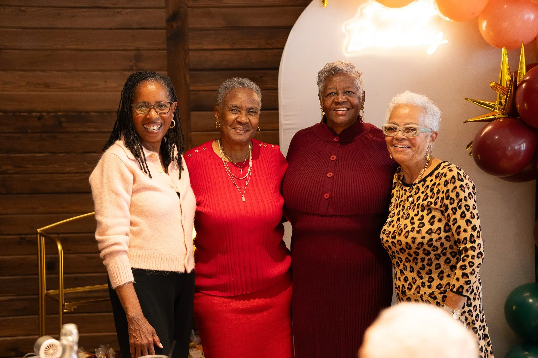 Four women standing side by side, smiling at the camera, at a celebration with balloons and wood-paneled wall in the background.