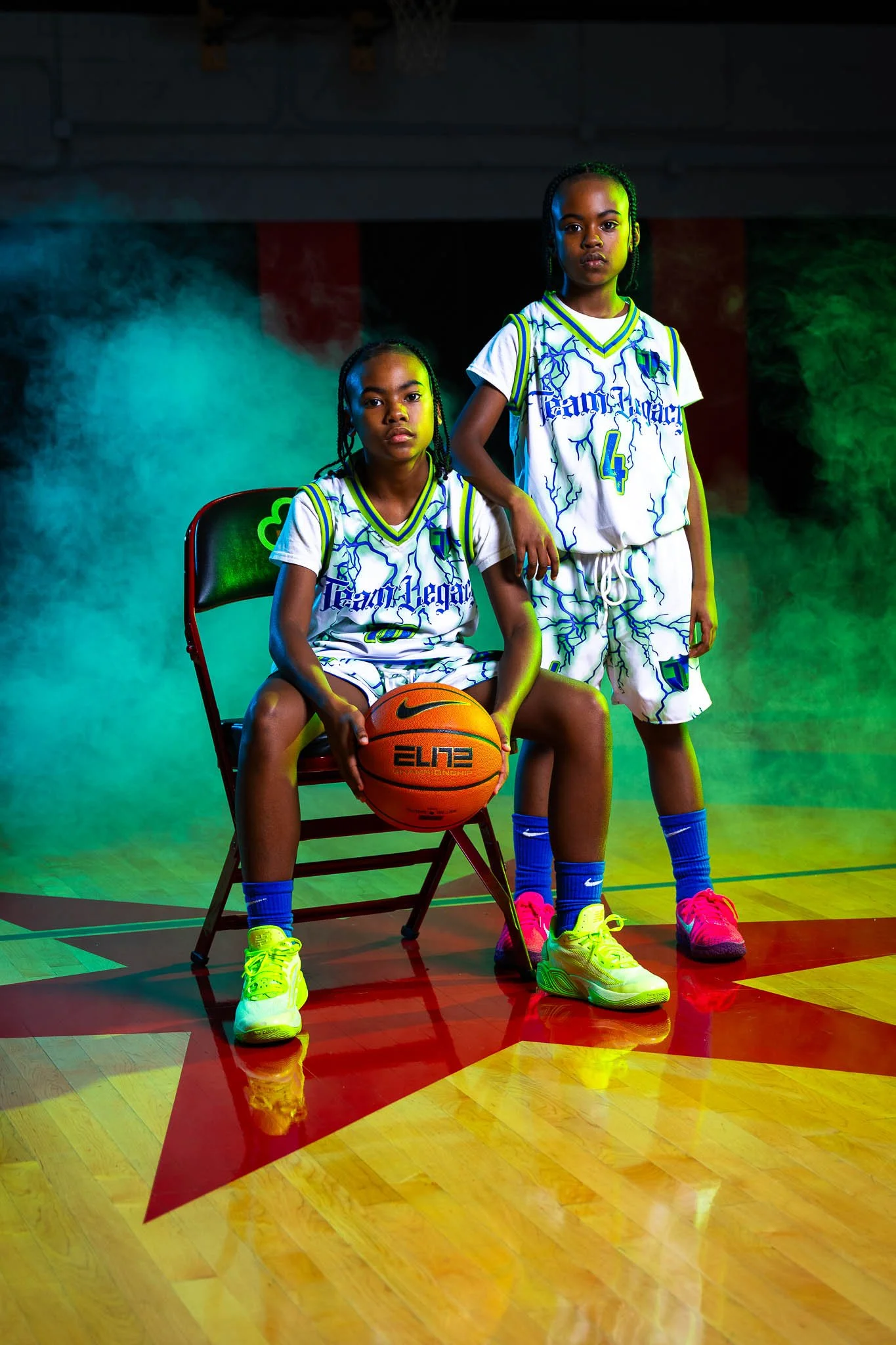 Two young girls in basketball uniforms, one sitting on a chair holding a basketball and the other standing beside her, in a gym with colorful lighting and fog atmosphere.