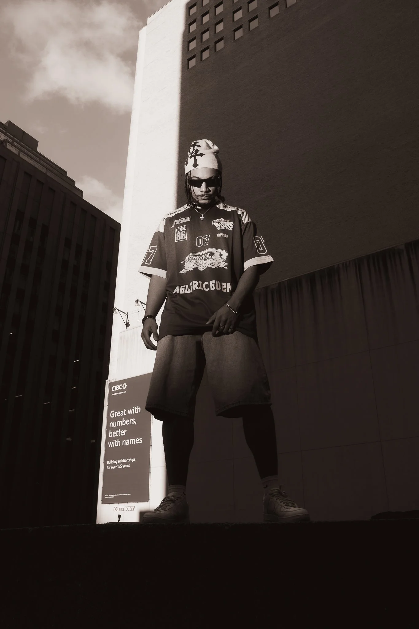 A person with a beanie, sunglasses, a sports jersey, shorts over leggings, and sneakers standing outdoors, with tall buildings and a billboard in the background.