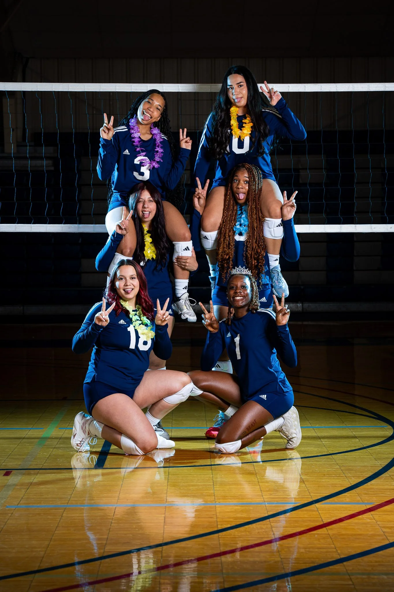 A group of six female volleyball players in blue uniforms posing on an indoor volleyball court. They are making peace signs and playful faces, with some sitting, kneeling, and standing in a pyramid formation. The net is visible in the background.