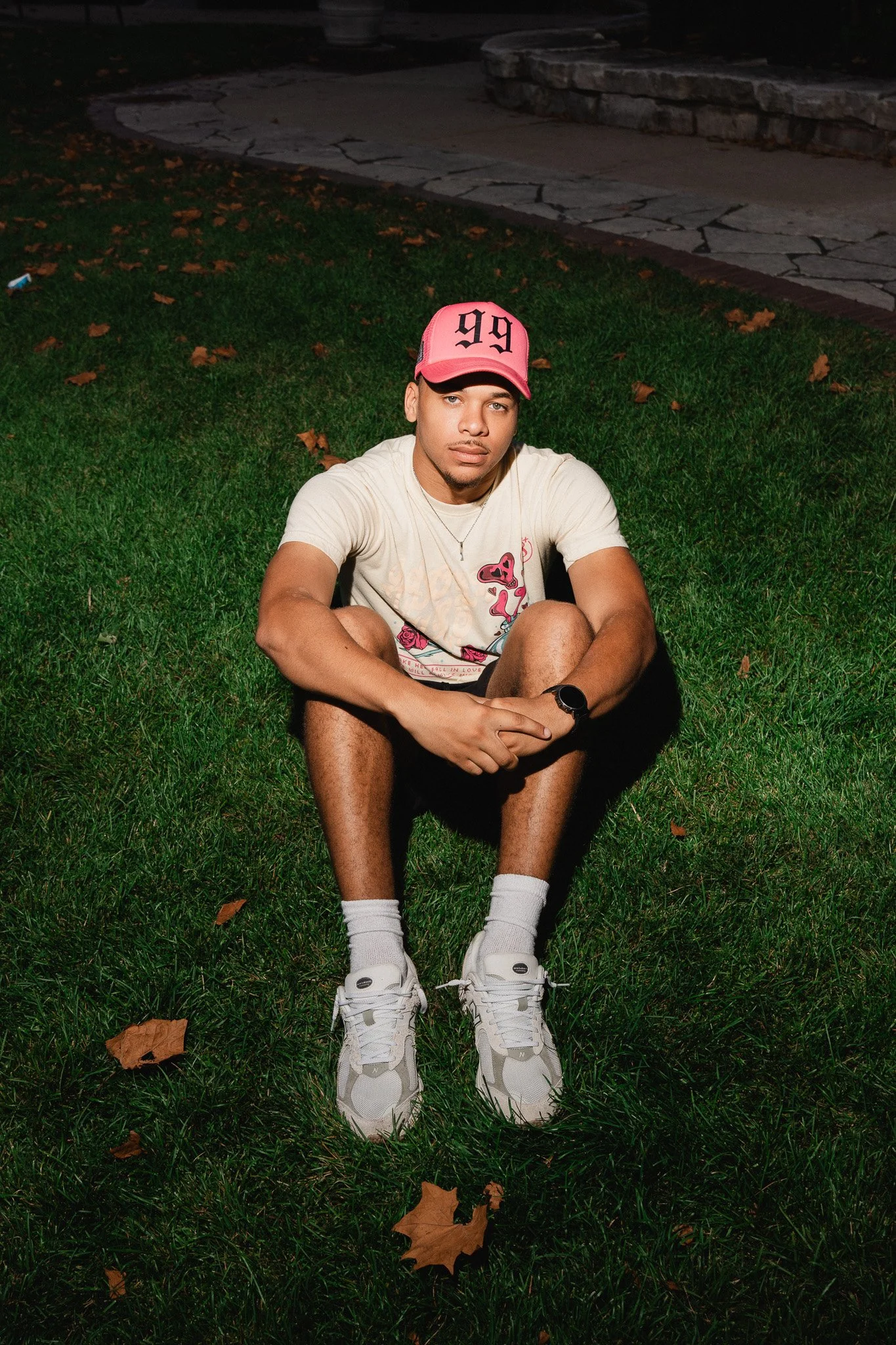 Young man sitting on grass at night, wearing a pink baseball cap, white graphic t-shirt, white sneakers, and black watch, with leaves scattered on the grass.