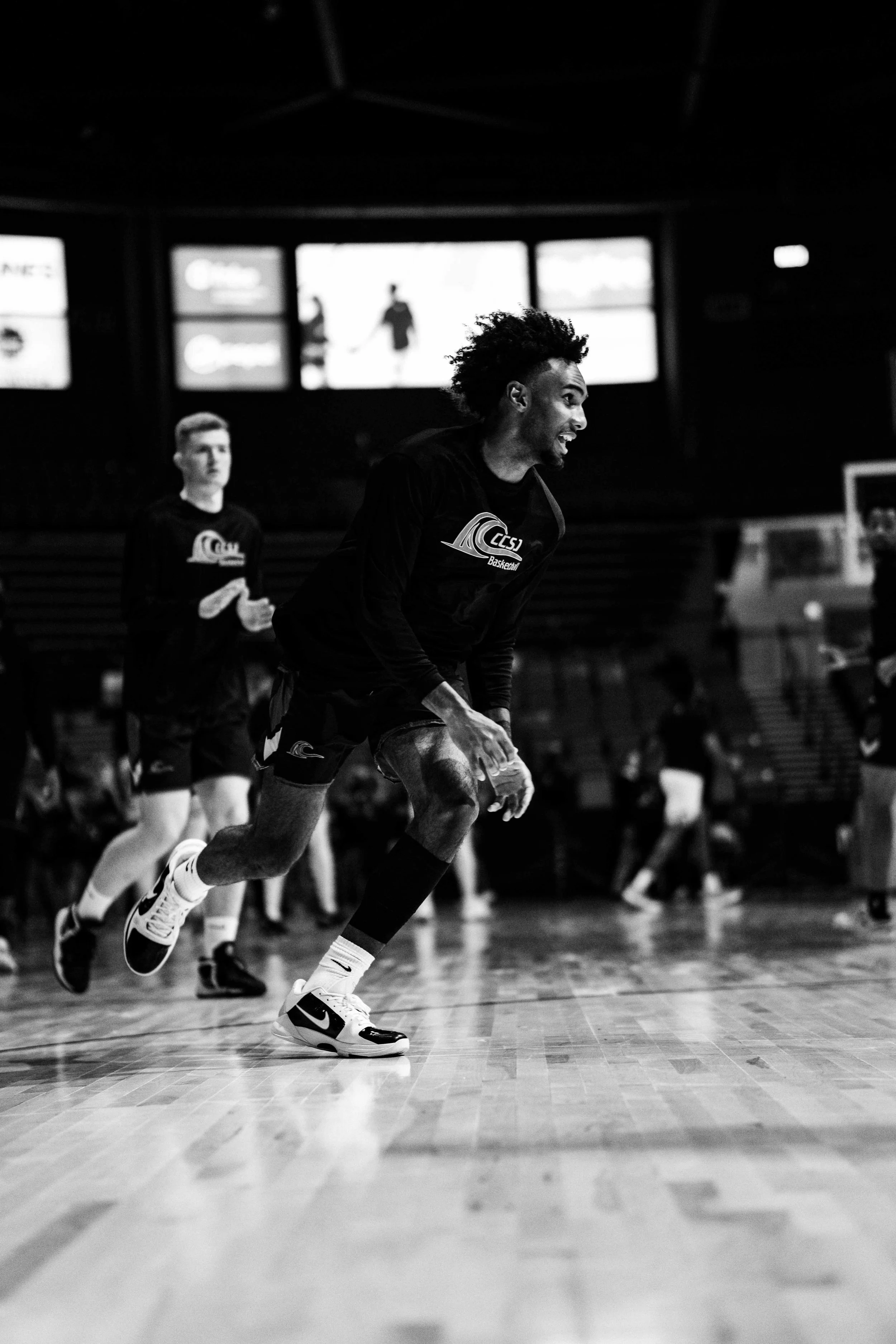 Black and white photo of a young man with curly hair running on an indoor basketball court, wearing a team jacket and athletic shoes, with other players in the background.
