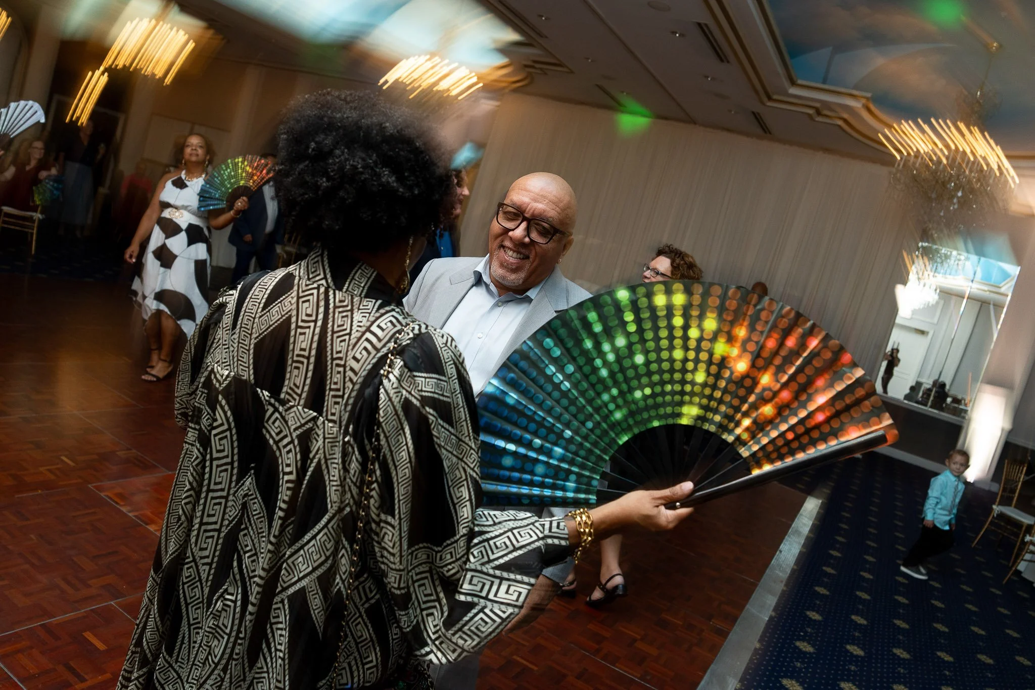 A woman with curly hair and an elegant black-and-white patterned dress is holding a colorful, illuminated fan and smiling at a man in a gray suit and glasses at a social event or celebration.