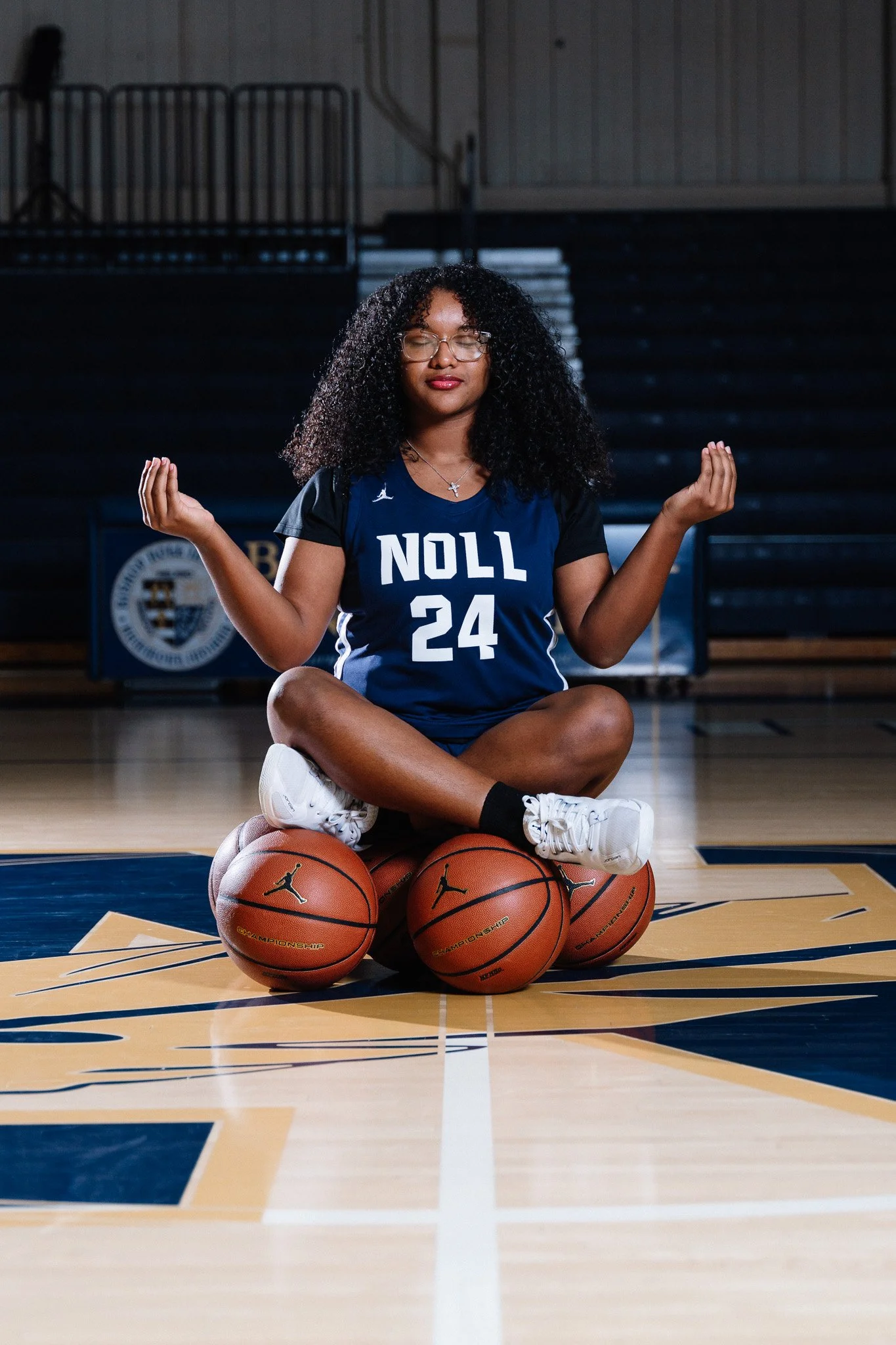 A woman with curly hair, wearing glasses and a basketball jersey with the text 'NOLL 24', is sitting cross-legged on a basketball court with three basketballs beneath her in a meditative pose.