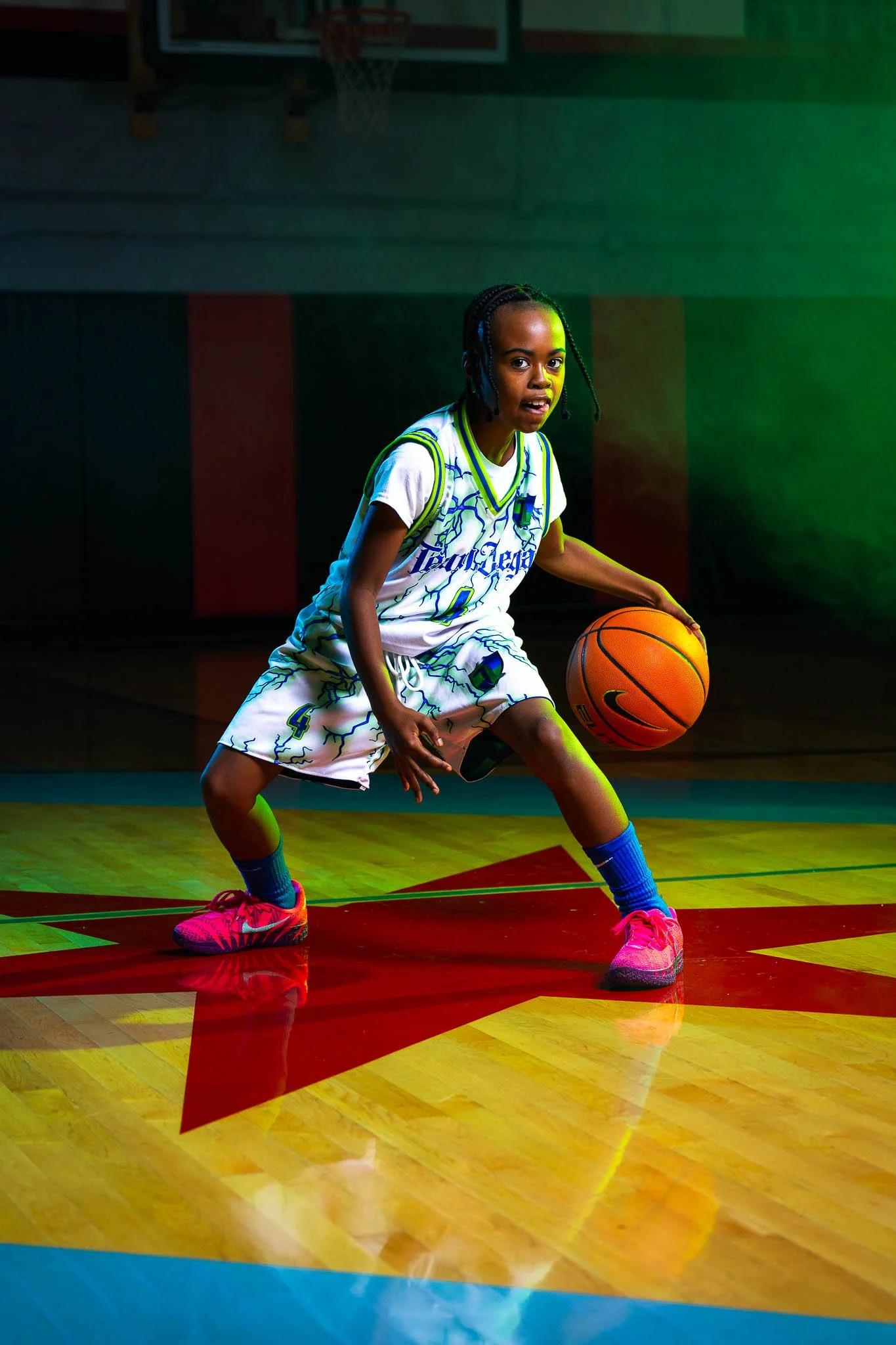 A young girl playing basketball on an indoor court, wearing a white basketball uniform with blue and green accents, pink sneakers, and blue socks, holding a basketball while crouching in a defensive stance.