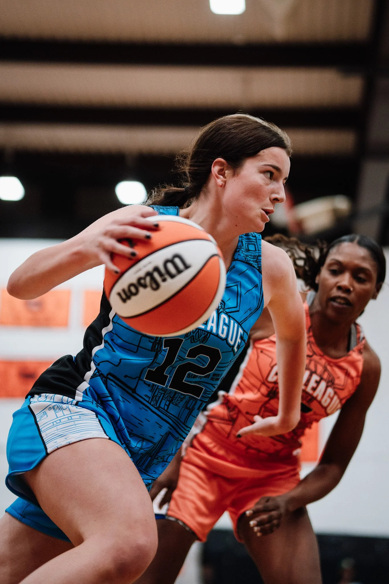 A female basketball player in a blue jersey holding a basketball, with a female opponent in an orange jersey defending behind her, during a game.