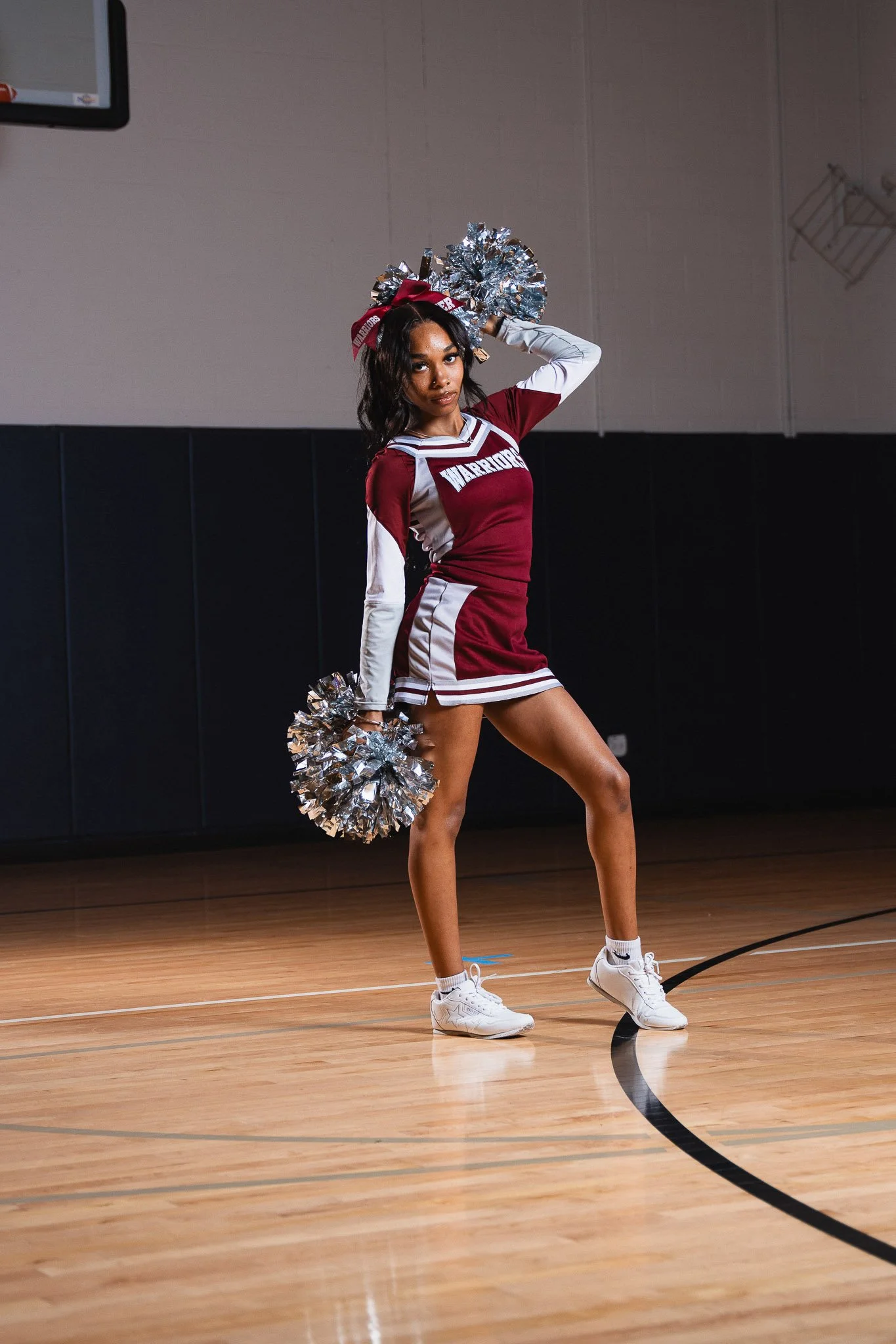 A cheerleader wearing a maroon and white uniform with 'WARriors' written on it, holding silver pom-poms and standing on a basketball court.