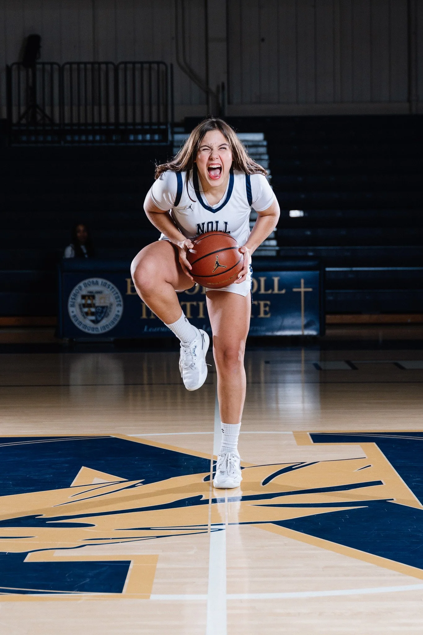 A female basketball player in a white jersey with blue and yellow accents, crouching and holding a basketball, on a basketball court with a logo at the center.