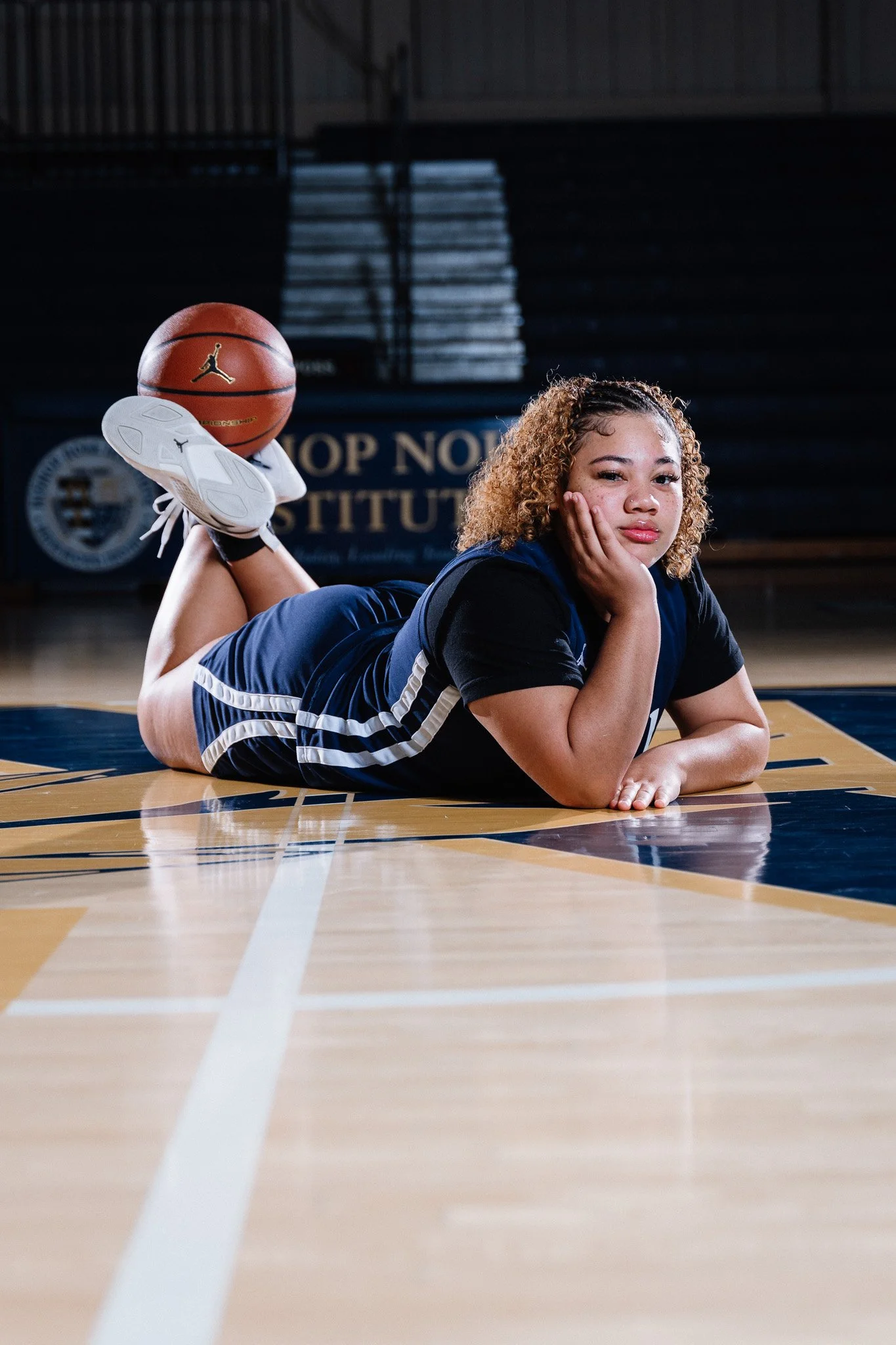 A young woman lies on her stomach on a basketball court, resting her head on her hand, with a basketball balanced on her feet.