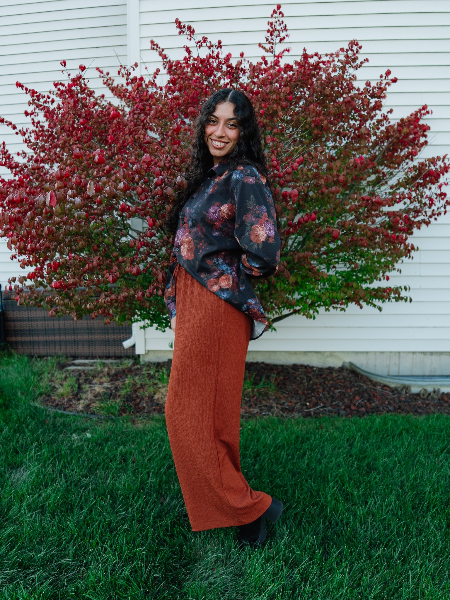 A woman with long, curly dark hair and a bright smile standing on green grass in front of a red and green bush, wearing a black floral blouse, rust-colored pants, and black shoes.