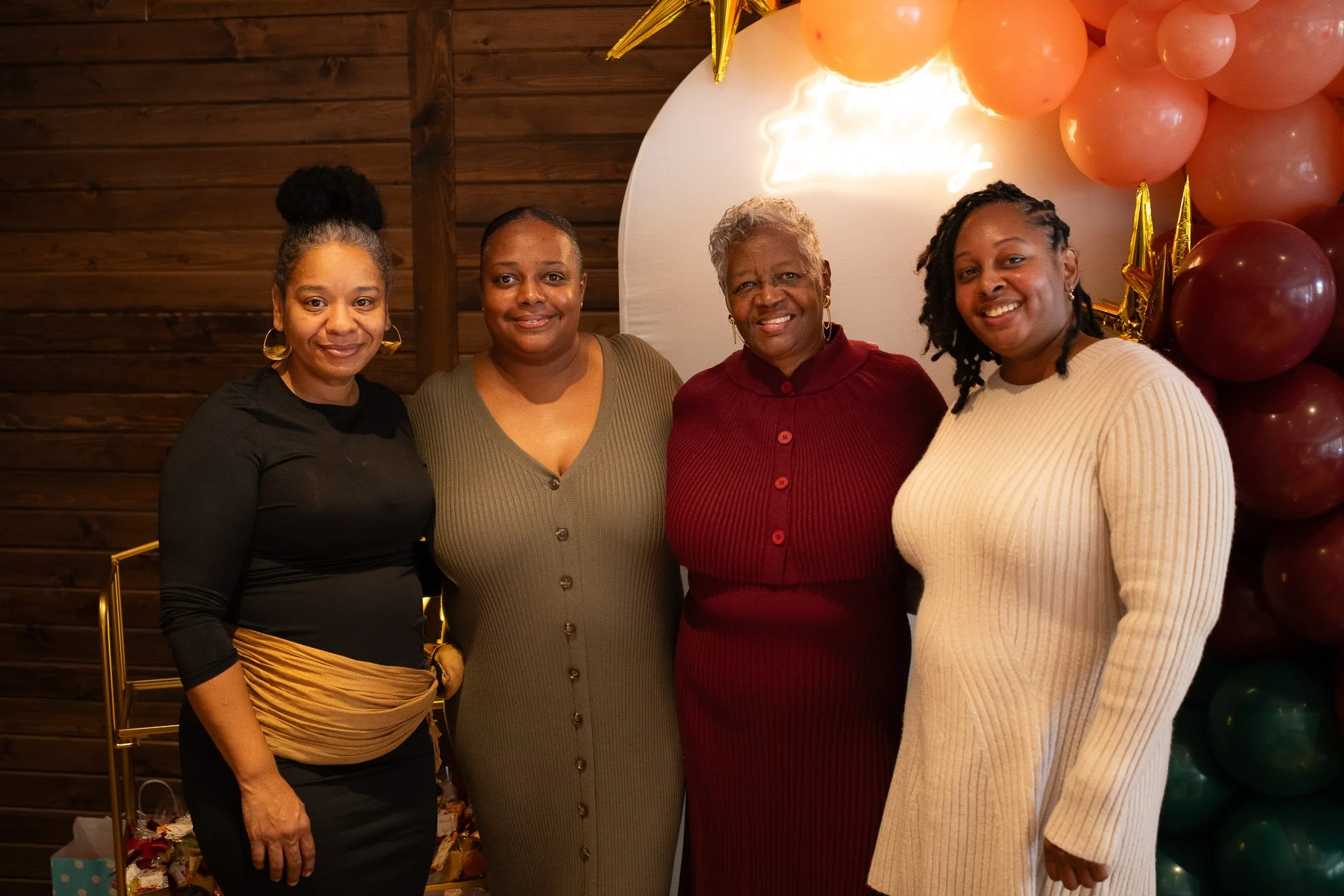 Four women standing together at a celebration, with balloons and a 'Happy Birthday' sign in the background.