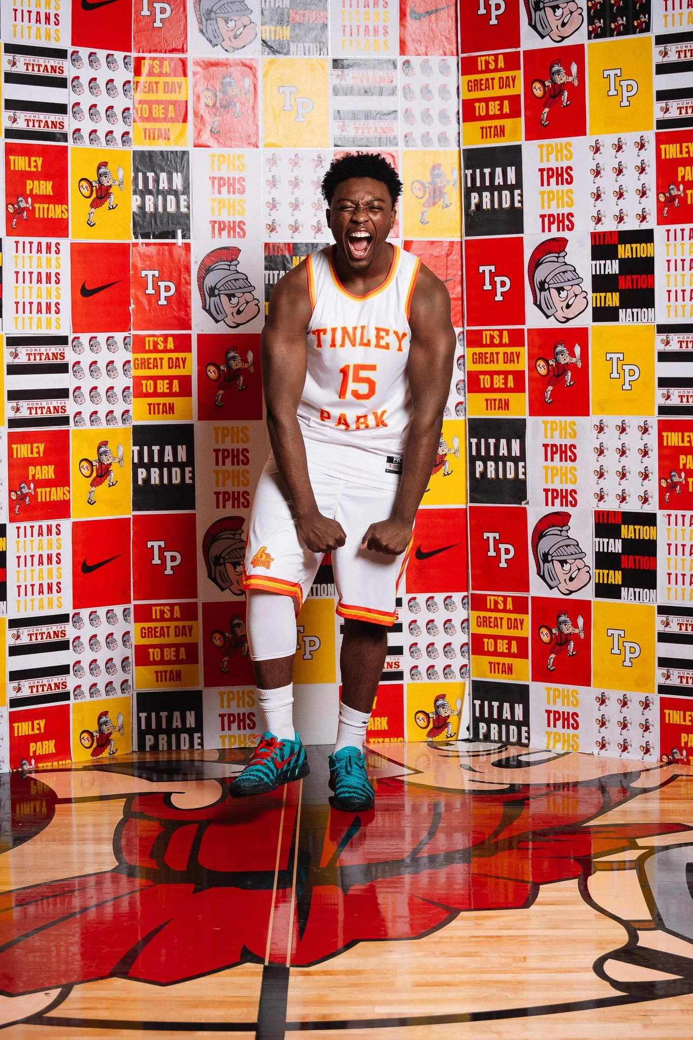 A young male basketball player wearing a white jersey with orange and yellow accents, number 15, standing on a basketball court with a smile and flexing his arms. The background features a colorful wall of posters and logos, including the words 'Tinl