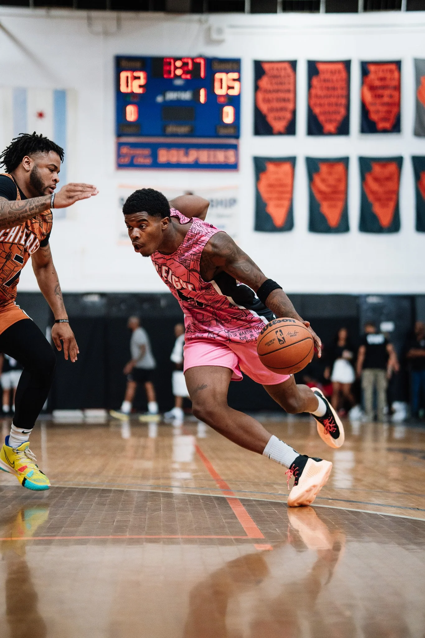 A basketball player in a pink uniform dribbling past an opponent in a black and orange uniform during a game. The game is taking place indoors with a scoreboard and banners visible in the background.