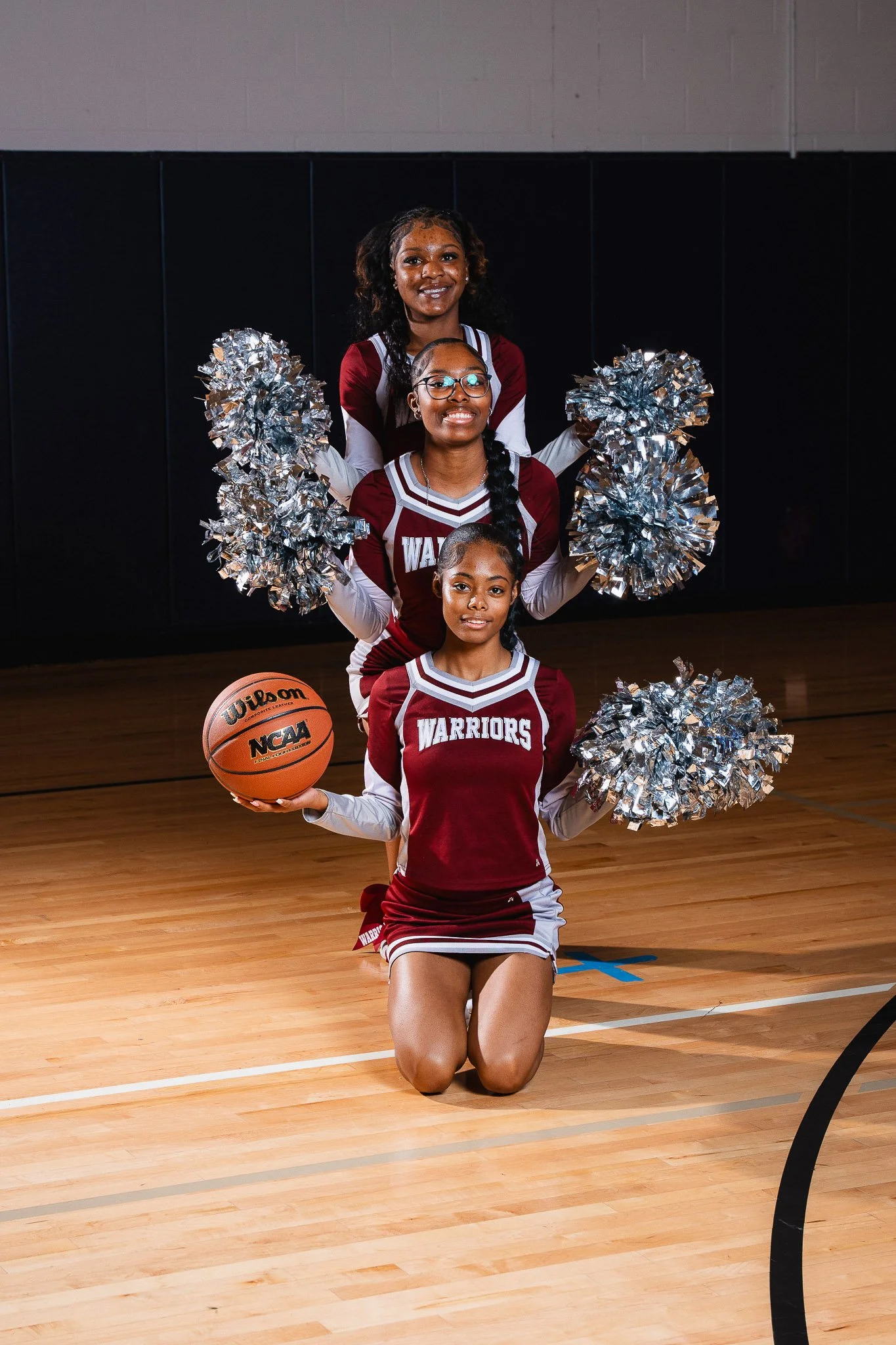 Three young female cheerleaders in maroon and white uniforms with 'WARRIORS' written on them, posing with silver pom-poms on a basketball court. The girl in the front is kneeling and holding a basketball with 'Wilson' and 'NCAA' written on it.