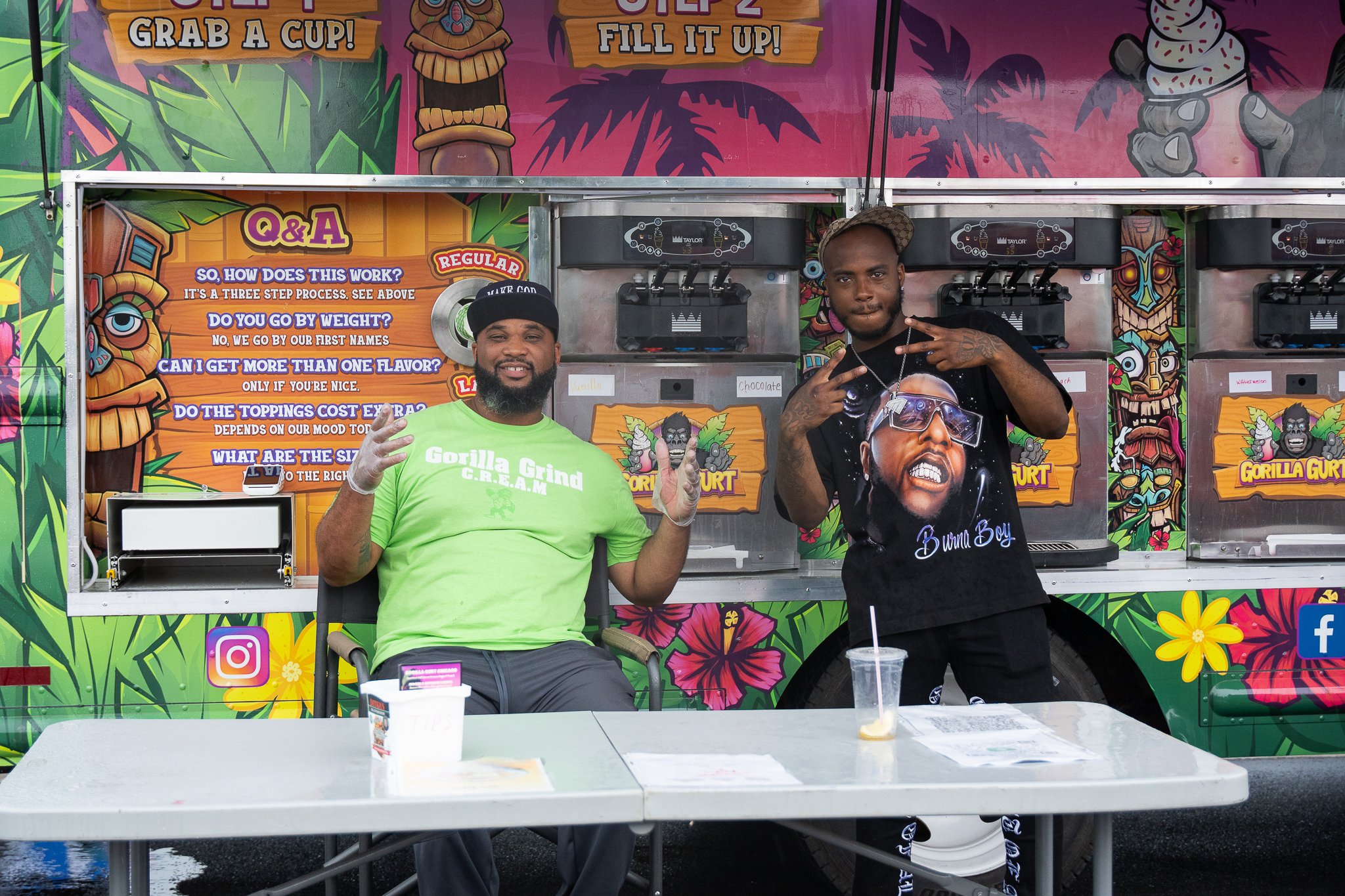 Two men posing in front of a colorful, tropical-themed Gorilla Gurt ice cream truck. One man, wearing a green Gorilla Grind shirt and a cap, is sitting and smiling with a peace sign. The other man, standing and making hand gestures, is wearing a blac