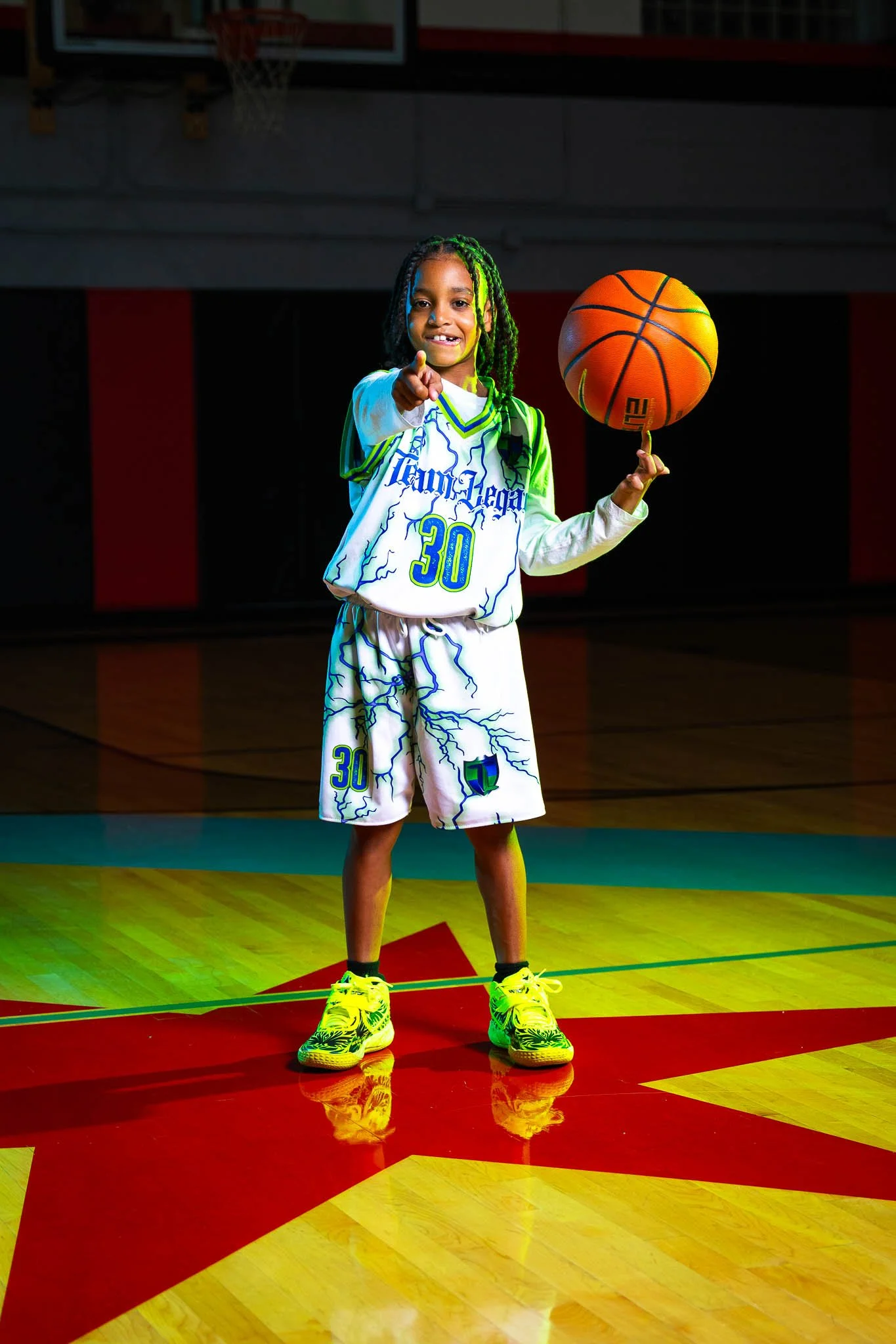A young girl standing on a basketball court, wearing a white basketball uniform with blue and green lightning bolt patterns and the number 30. She is holding an orange basketball and pointing directly at the camera, smiling. She has dark braided hair