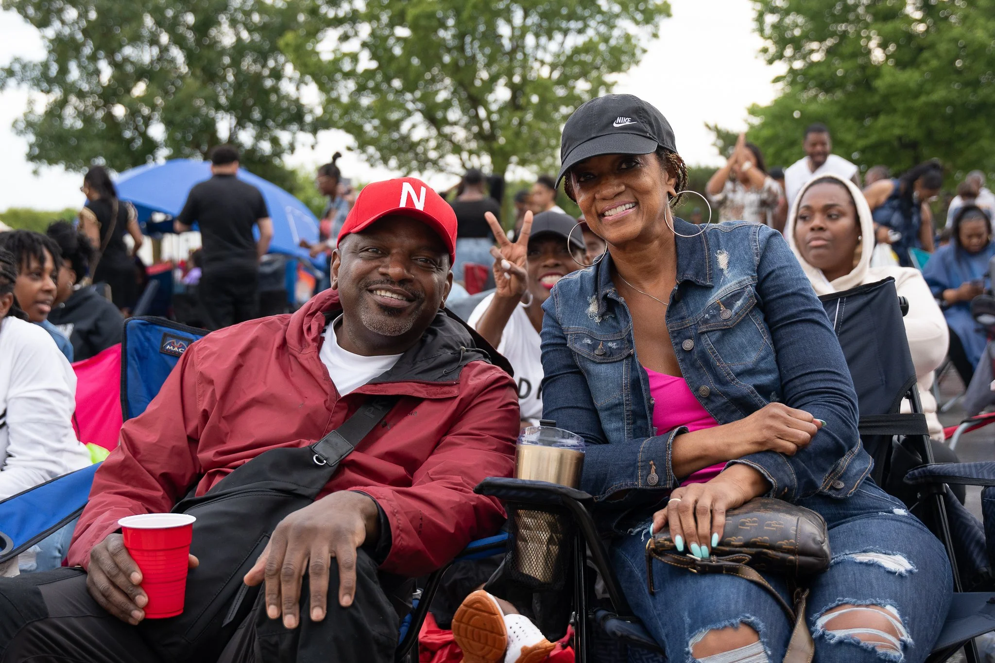 Smiling man wearing a red cap and red jacket sitting next to a smiling woman wearing a black cap and denim jacket at an outdoor event surrounded by a crowd of people.