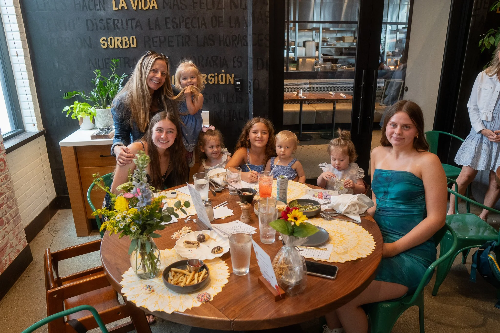 A group of women and children sitting and standing around a table in a restaurant, celebrating. The table has snacks, drinks, and flowers. The background shows a black wall with gold lettering and a view into the kitchen.