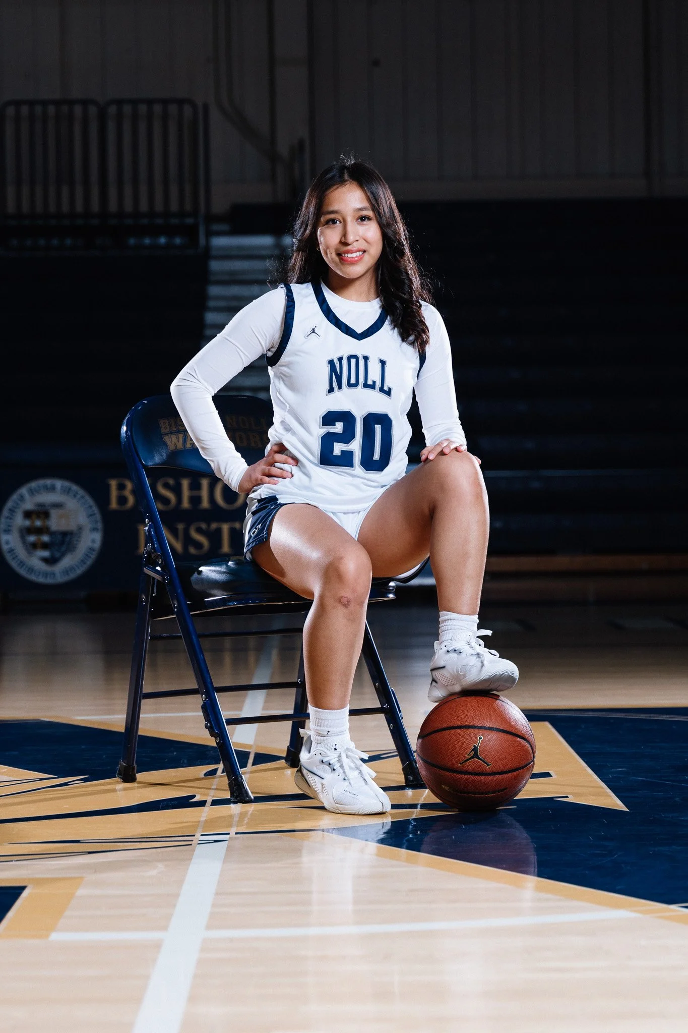 A young woman in a basketball uniform sitting on a chair on a basketball court, with her right hand on her hip and her left leg resting on a basketball.