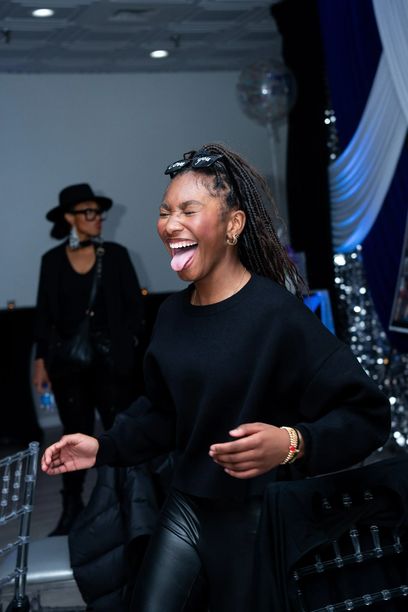 A young woman with braided hair and sunglasses on her head, smiling and sticking out her tongue at a party or celebration.