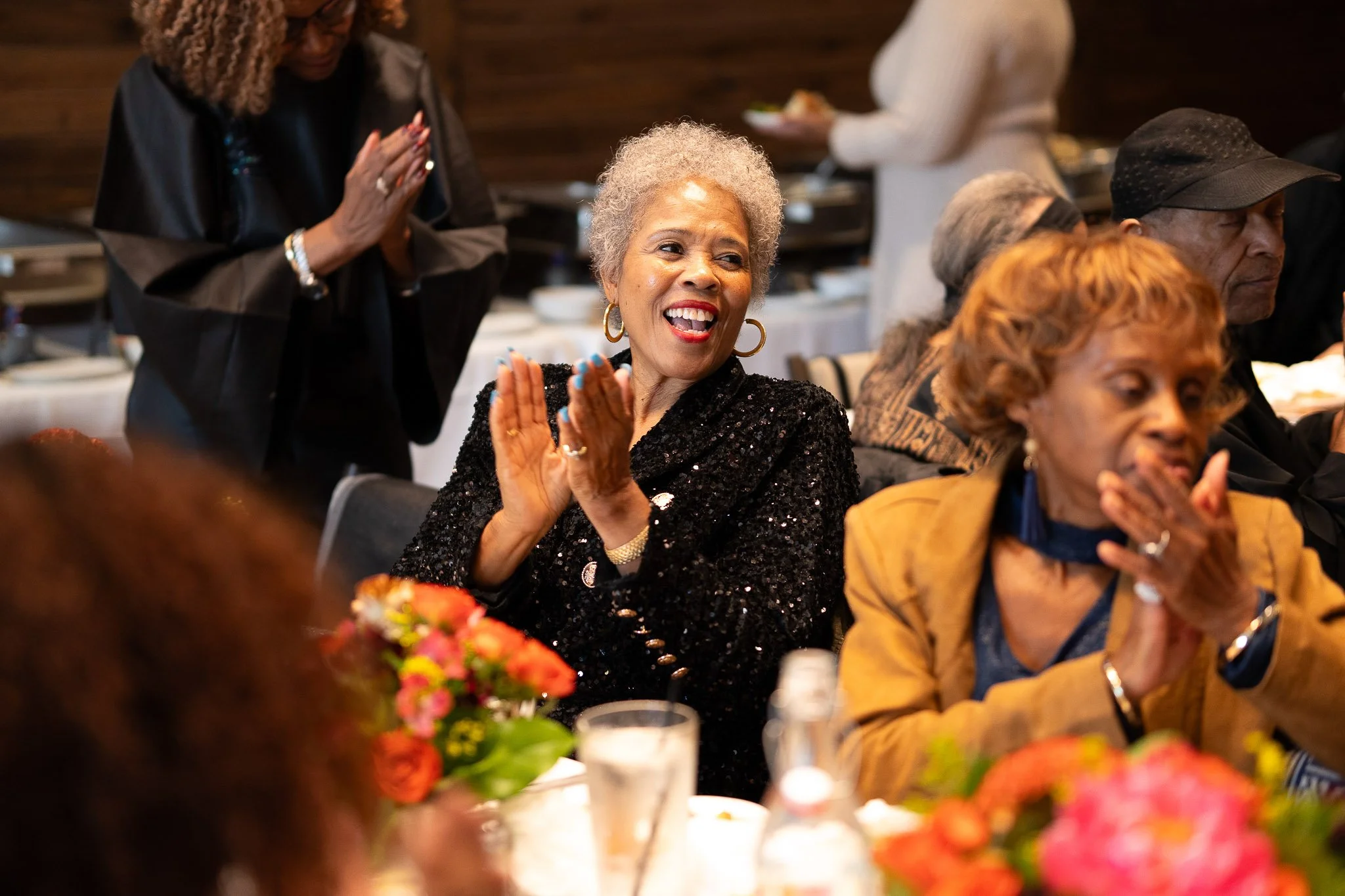 A joyful elderly woman with short gray hair, wearing a black sequined jacket, is clapping and smiling at a gathering or celebration. She is seated at a table decorated with pink and orange flowers.