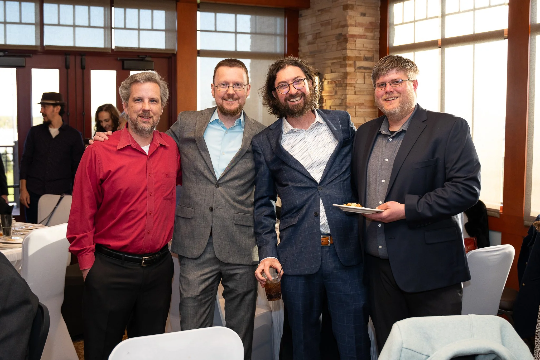 Four men in business attire smiling and posing for a photo at a social event in a well-lit room. Two of them are holding drinks, and one man is holding a plate of food.