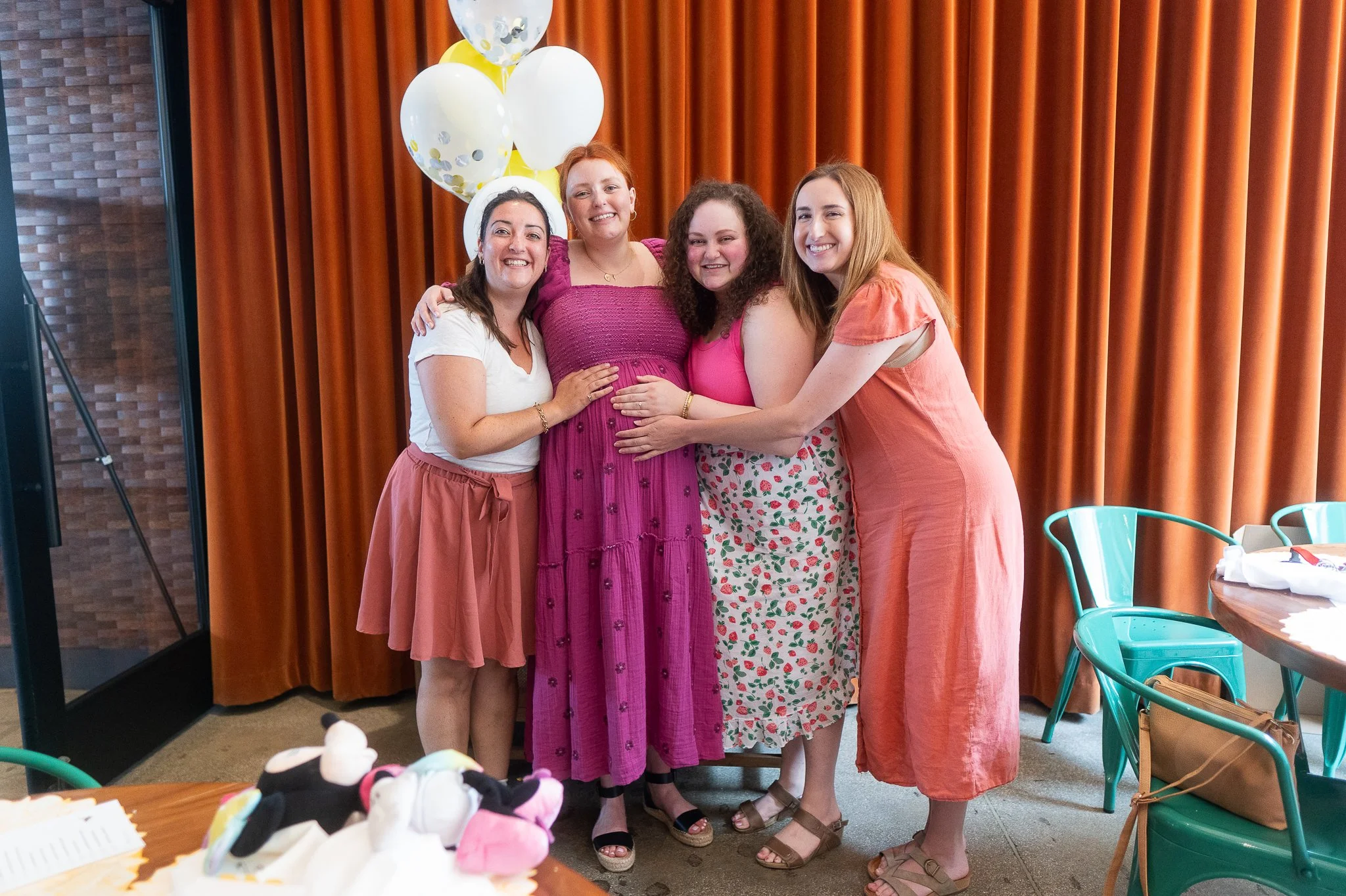 Group of four women smiling and hugging, standing in front of a brown curtain, with balloons behind them, at a celebration or gathering.