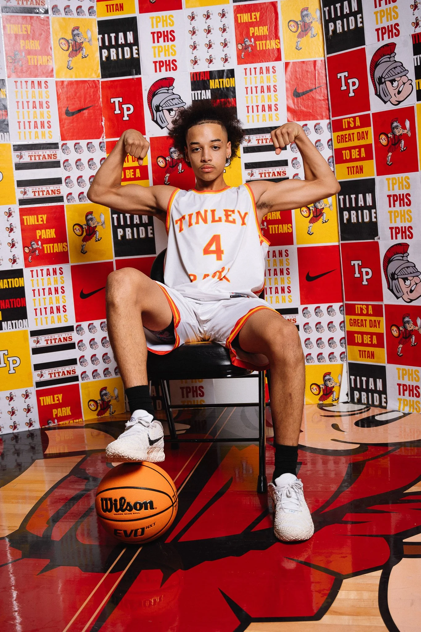 A young female basketball player sitting on a chair in a gymnasium, flexing her arms, wearing a white basketball uniform with orange and yellow accents, number 4, with a Wilson basketball on the floor in front of her. The background is decorated with