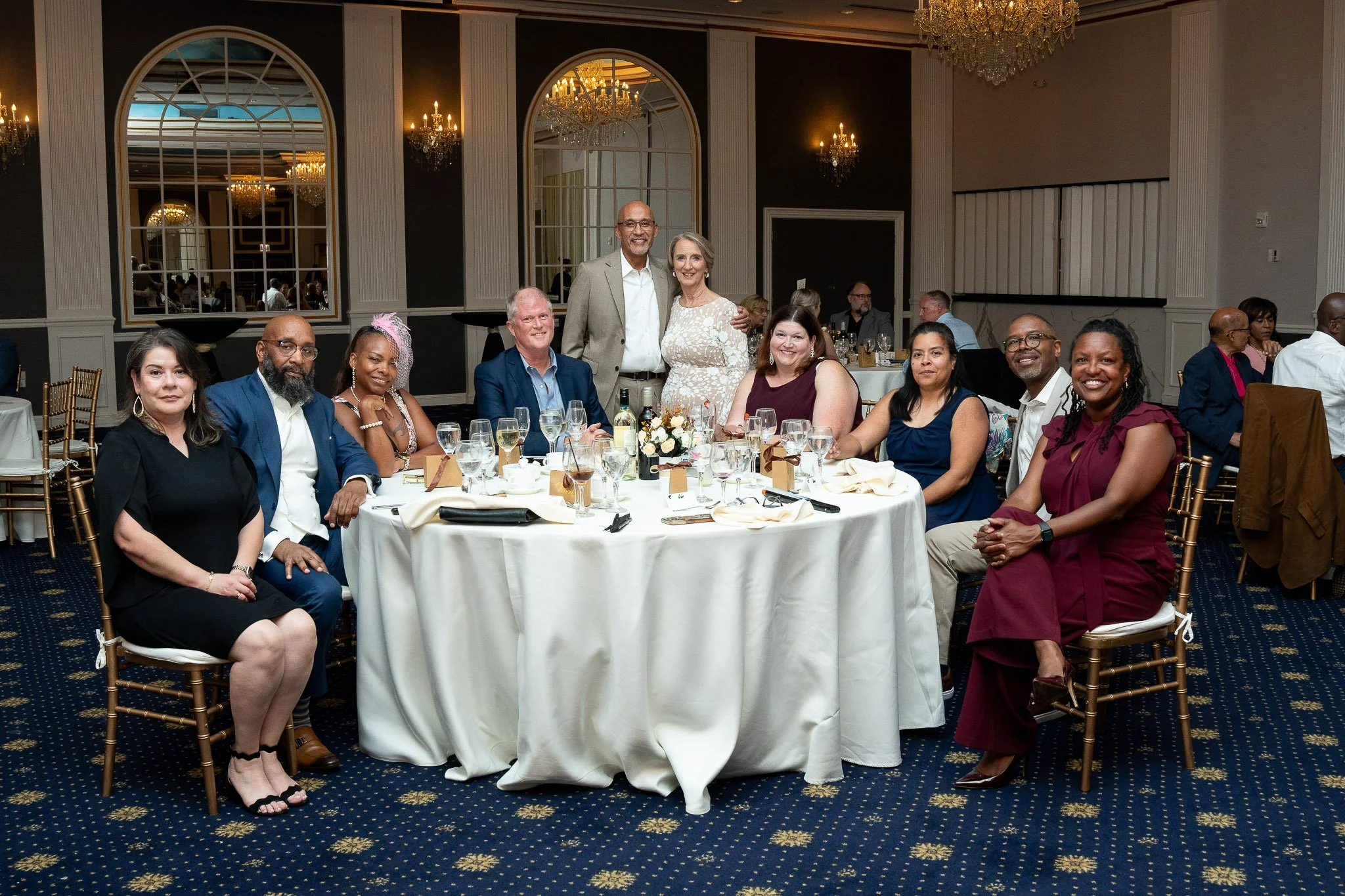 Group of eleven diverse adults sitting and standing around a banquet table at a formal event in an elegant banquet hall with chandeliers and large mirrors.