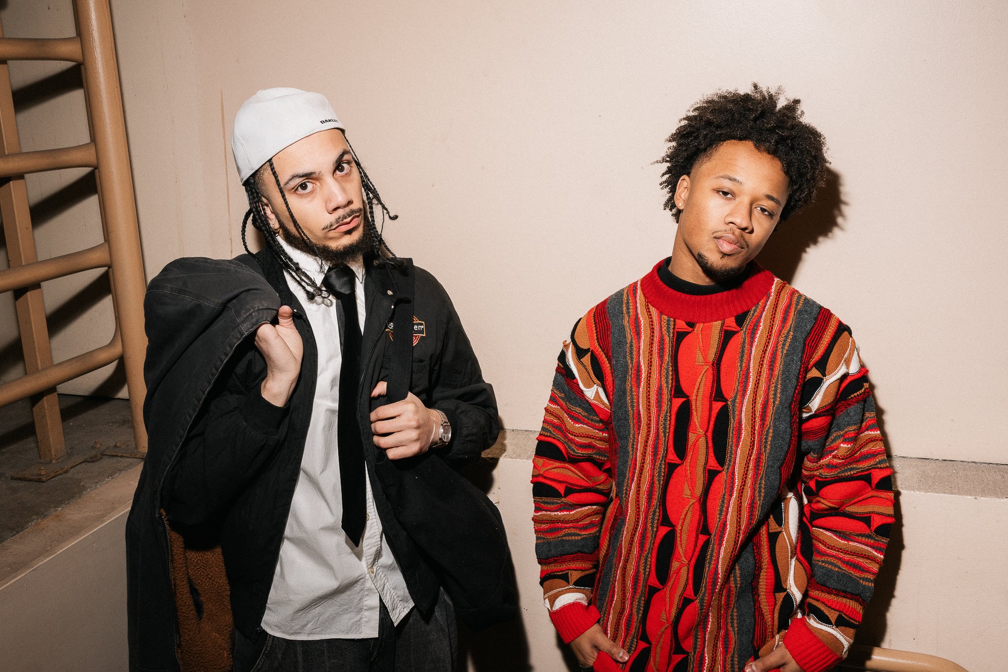 Two young men standing against a plain wall, one on the left wearing a white cap, black jacket, and white shirt, and the other on the right wearing a colorful patterned sweater.