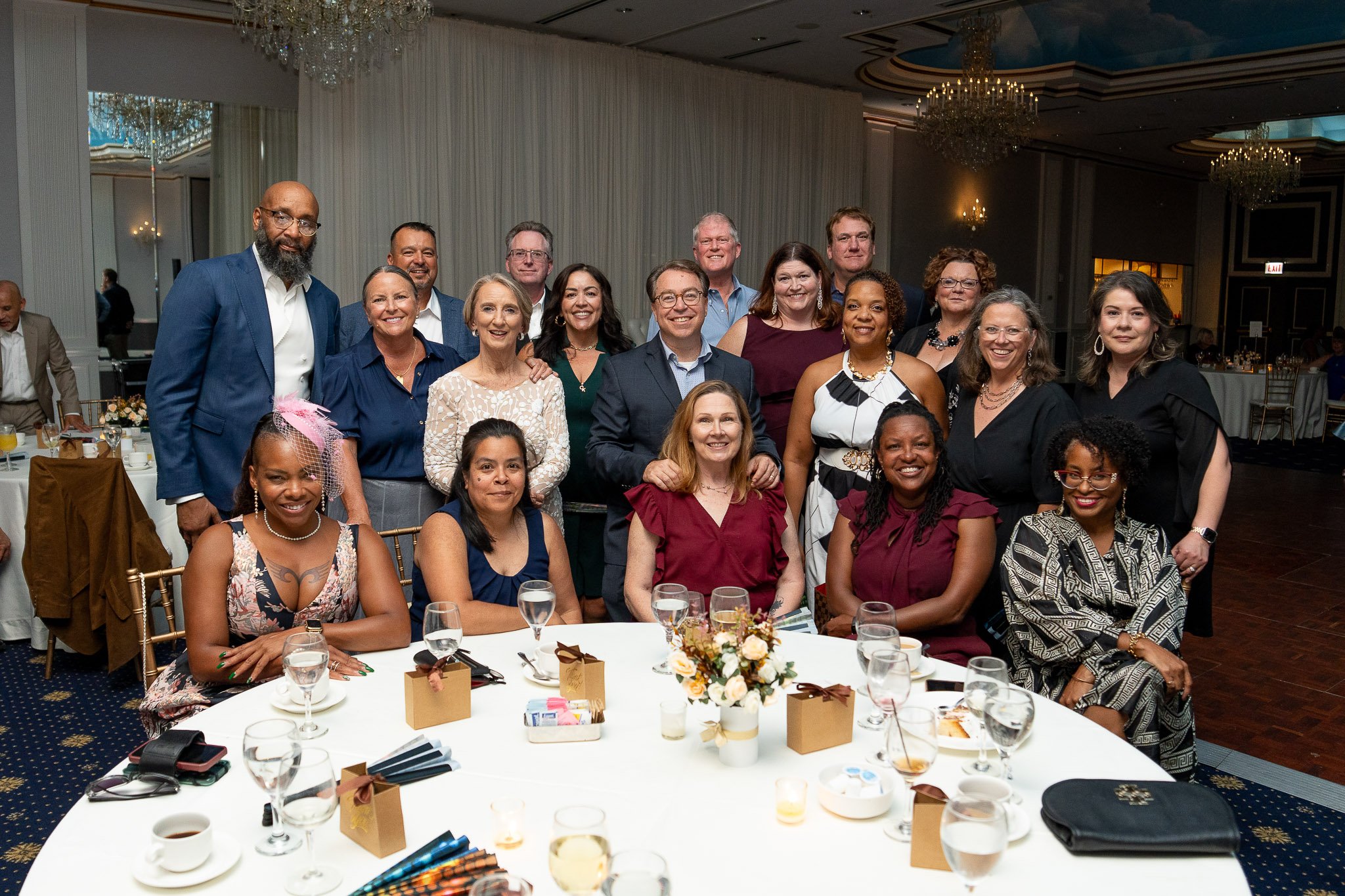 A group of 17 people, mostly women, gathered around a table at a formal event in a banquet hall. The table has floral centerpiece, drinks, and gift boxes, with elegant decorations and chandeliers in the background.