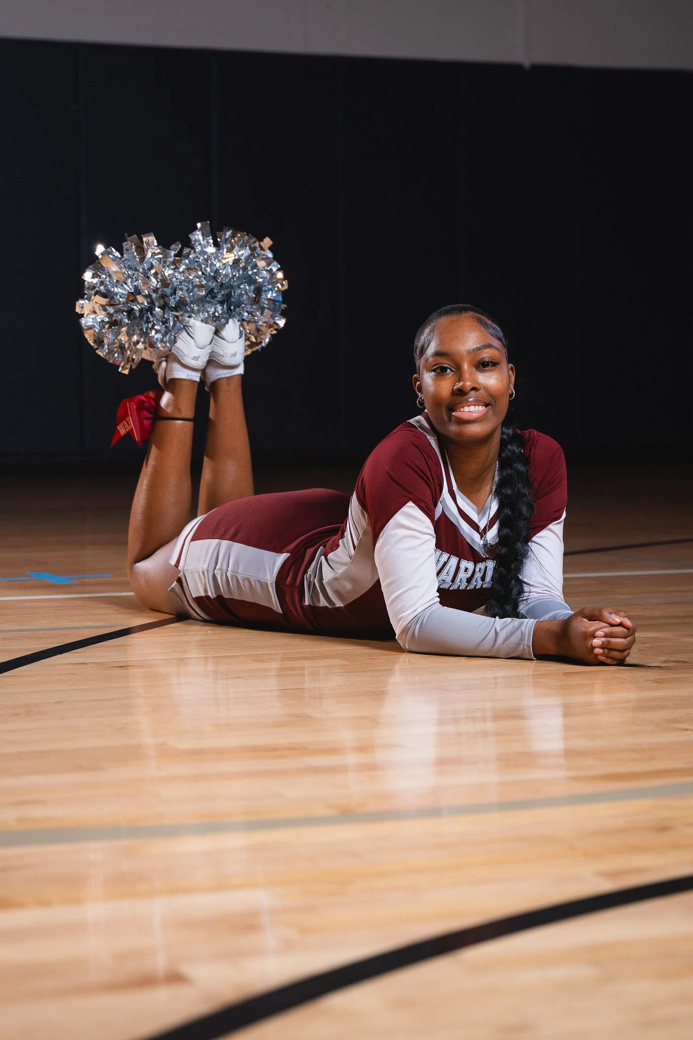 A young woman in a maroon and white cheerleading uniform lying on her stomach on a basketball court, holding silver pom-poms and smiling at the camera.