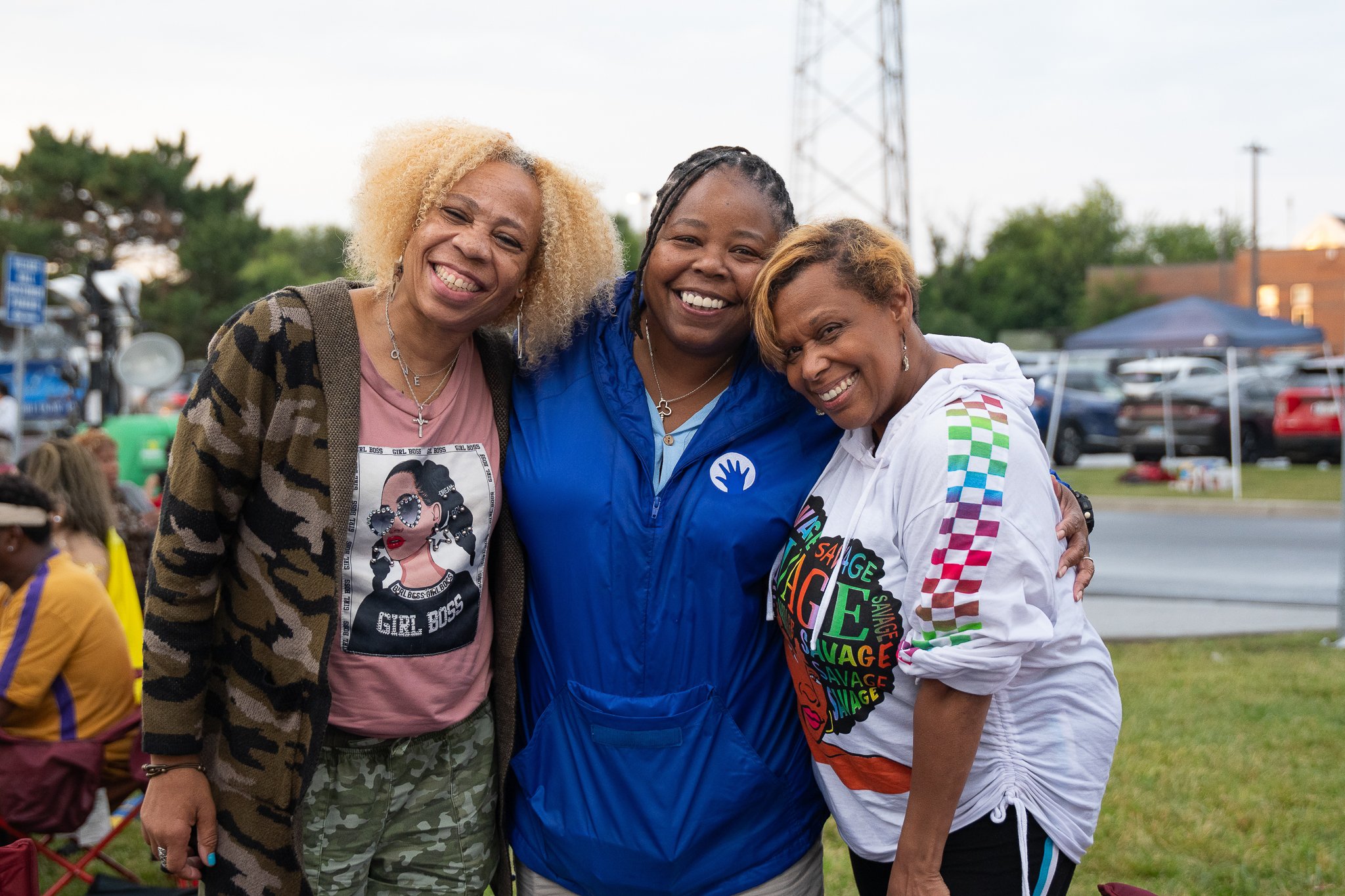 Three women smiling and hugging outdoors at a gathering or event, with cars and a tent in the background.