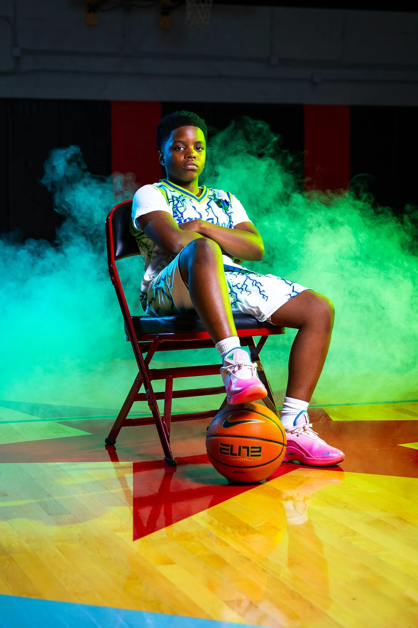 A young girl sitting on a red folding chair on a basketball court. She is wearing a white and blue sports outfit, pink basketball shoes, and has a basketball placed under her right foot. She is crossing her arms and looking confidently at the camera 