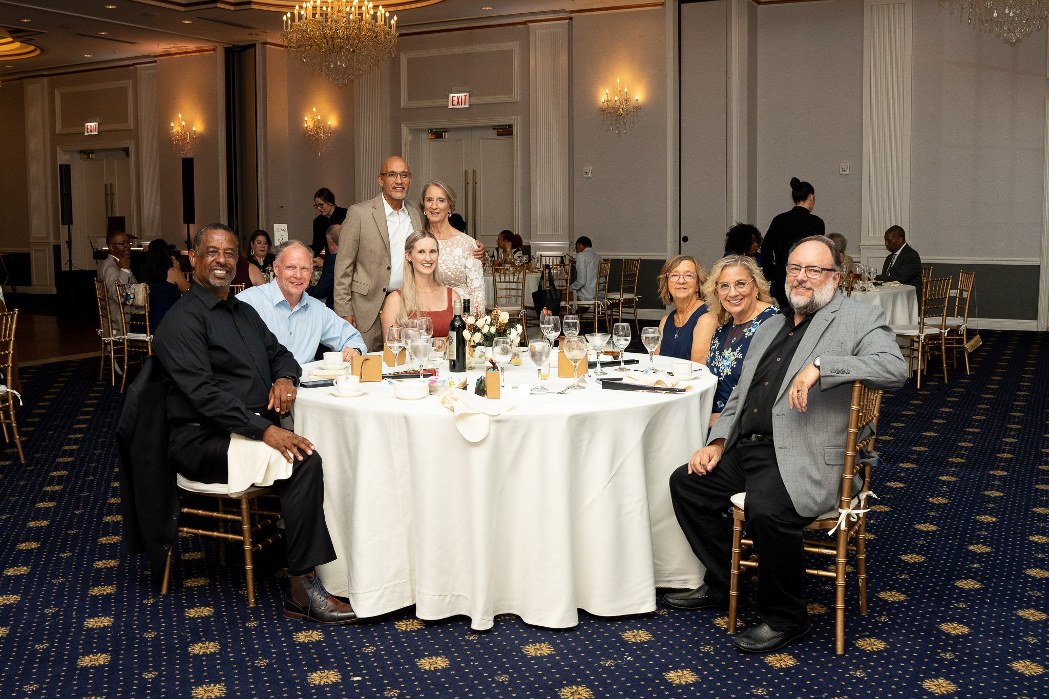 A group of nine people sitting and standing around a round table at a formal event in a ballroom, smiling at the camera. The table is decorated with wine bottles, glasses, and floral arrangements.