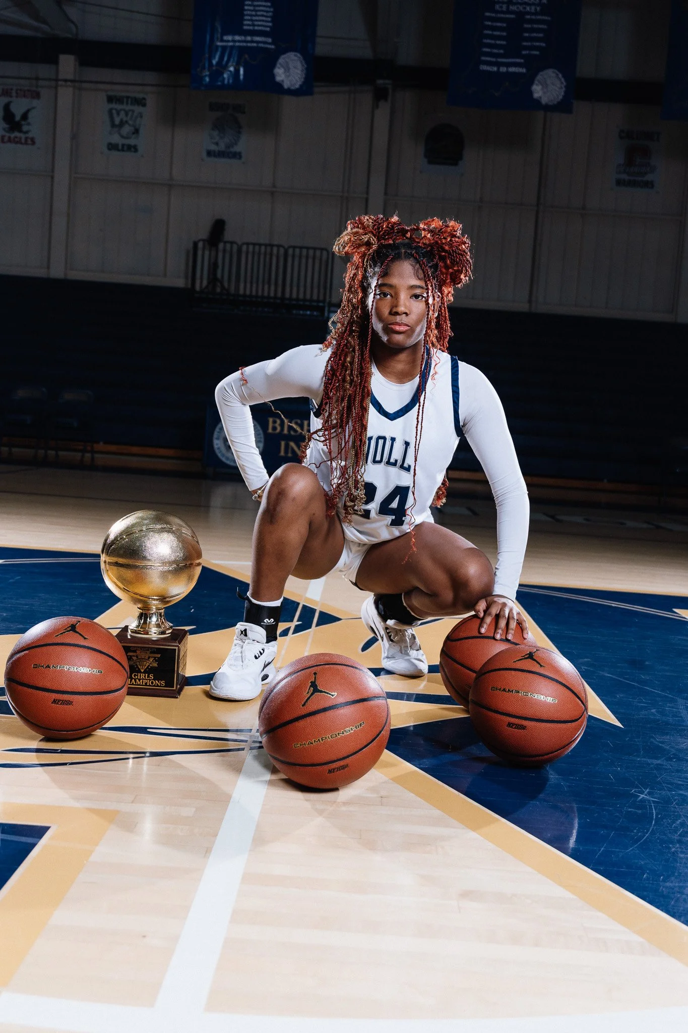 Young female basketball player wearing a white and blue uniform with the number 24, crouching on a basketball court with three basketballs and a trophy in front of her, inside a gymnasium with banners hanging in the background.