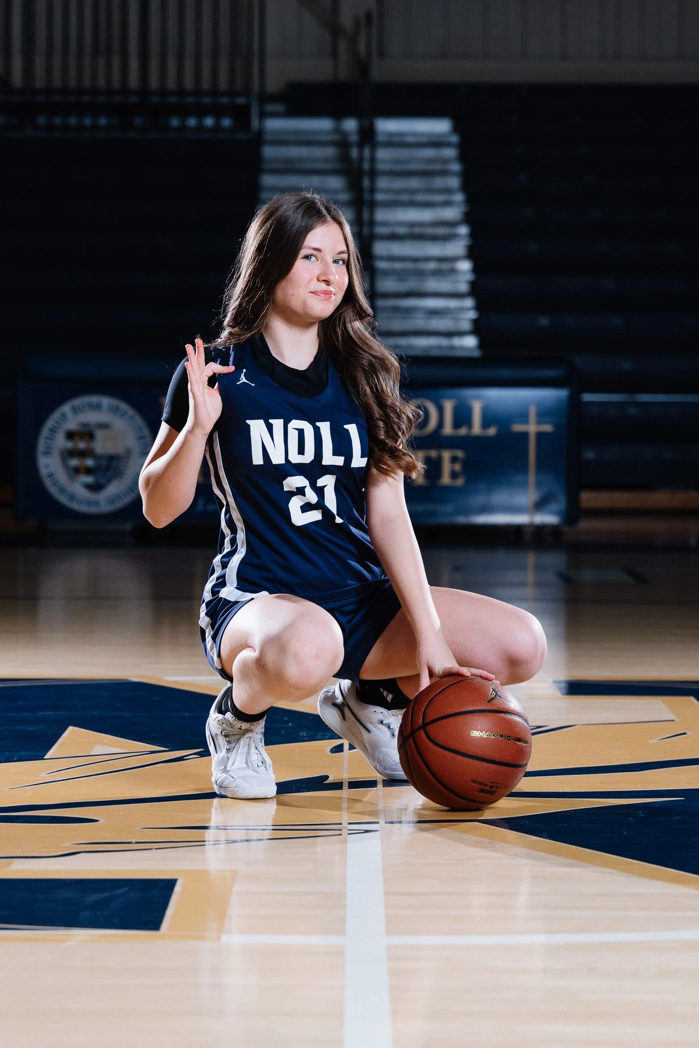 A young woman in a blue basketball uniform kneels on a basketball court, holding a basketball with the university logo visually on it, making the okay hand gesture with her right hand.
