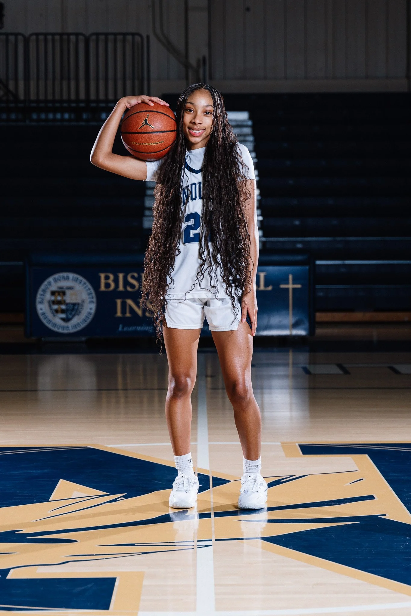 A young female basketball player holding a basketball on her shoulder, standing on a basketball court inside a gymnasium.